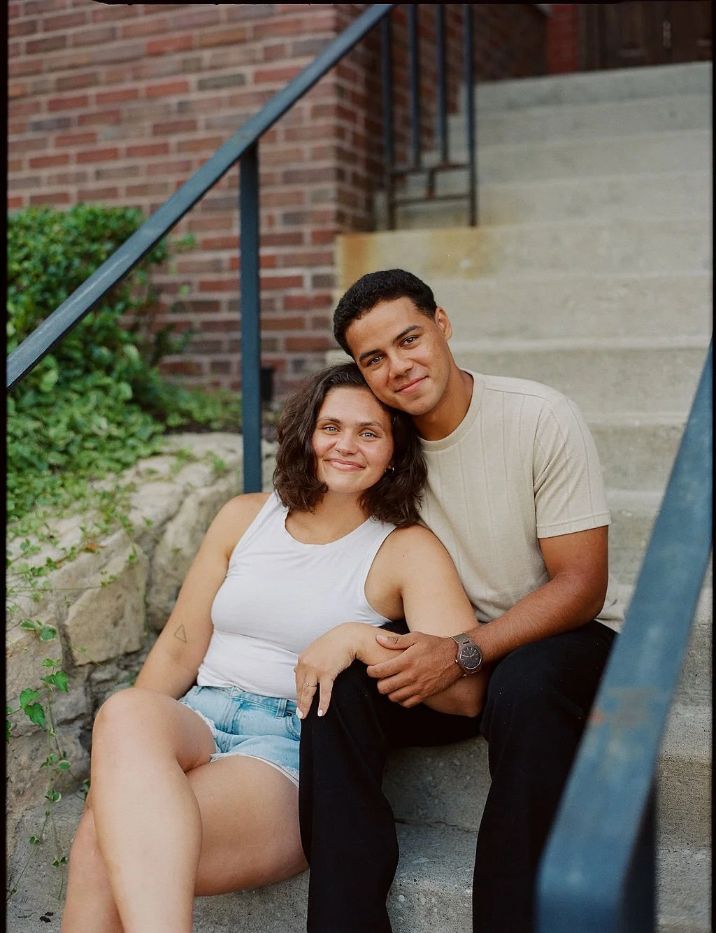A smiling couple sitting on concrete stairs outdoors, with the woman leaning against the man, surrounded by green plants and a brick building.