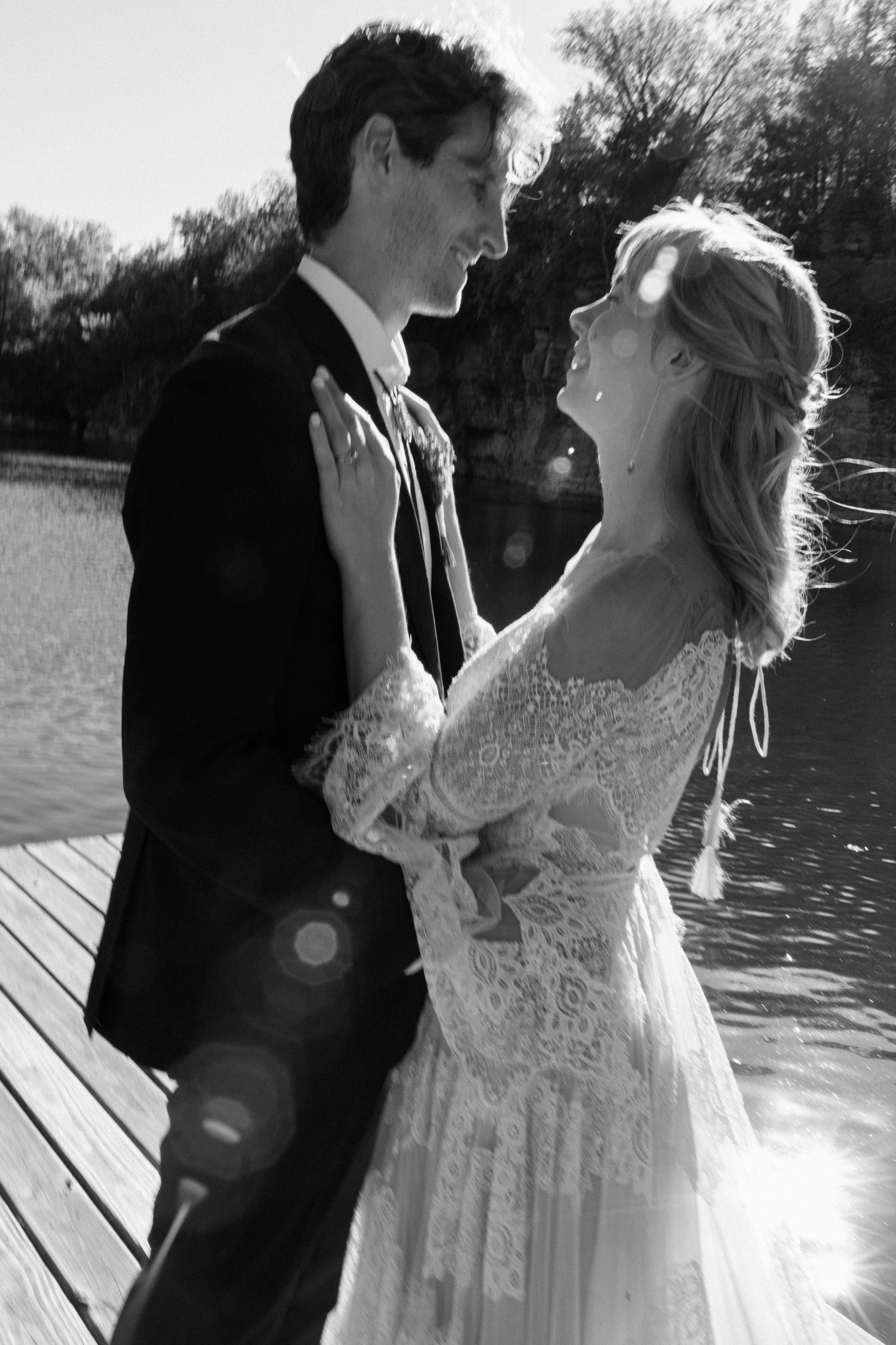 A black and white photo of a bride and groom smiling and looking at each other by a lake, with trees in the background and sunlight creating lens flares.