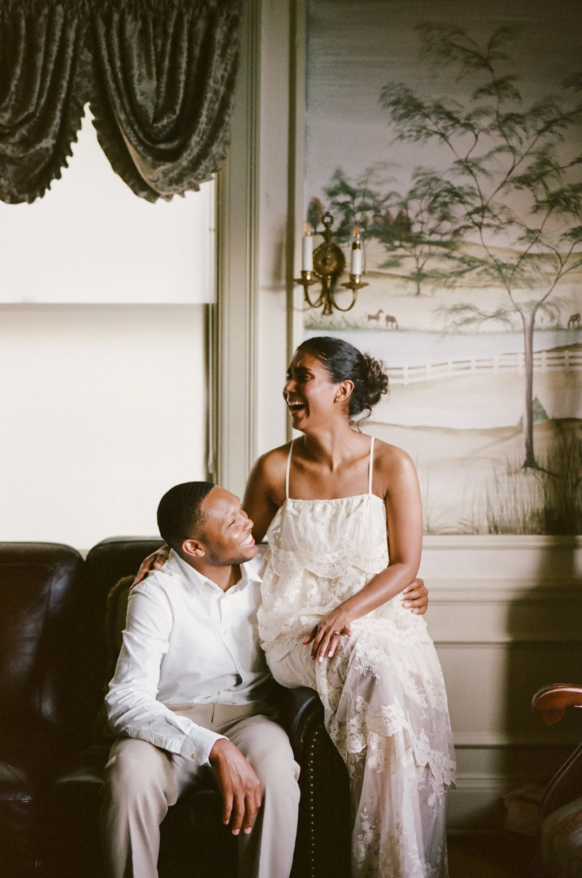 A man and woman sitting on a dark brown leather couch in a living room, smiling and laughing together.