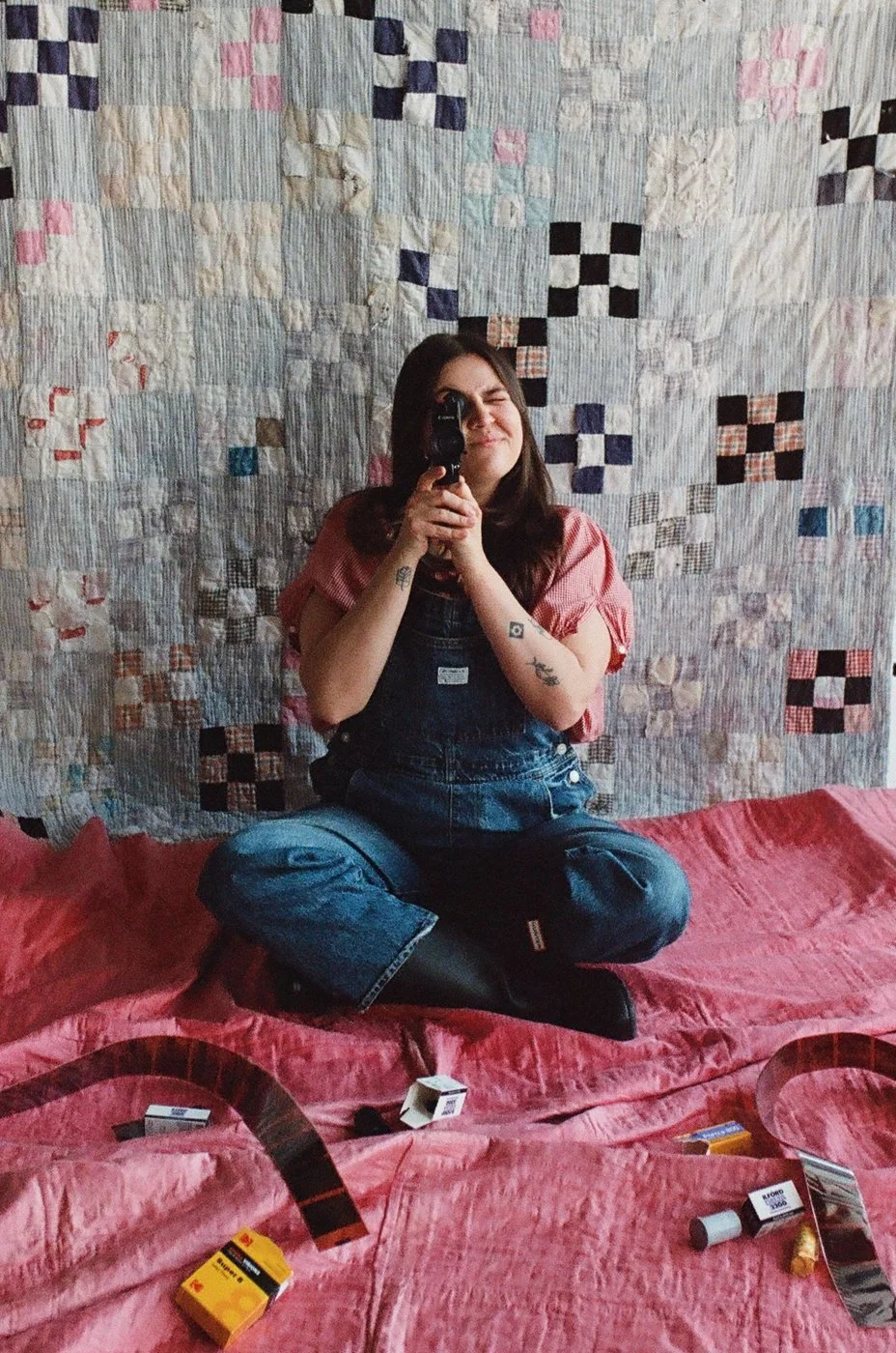 A woman sitting cross-legged on a pink bedspread, with a Super 8 camera in front of a hanging quilt.