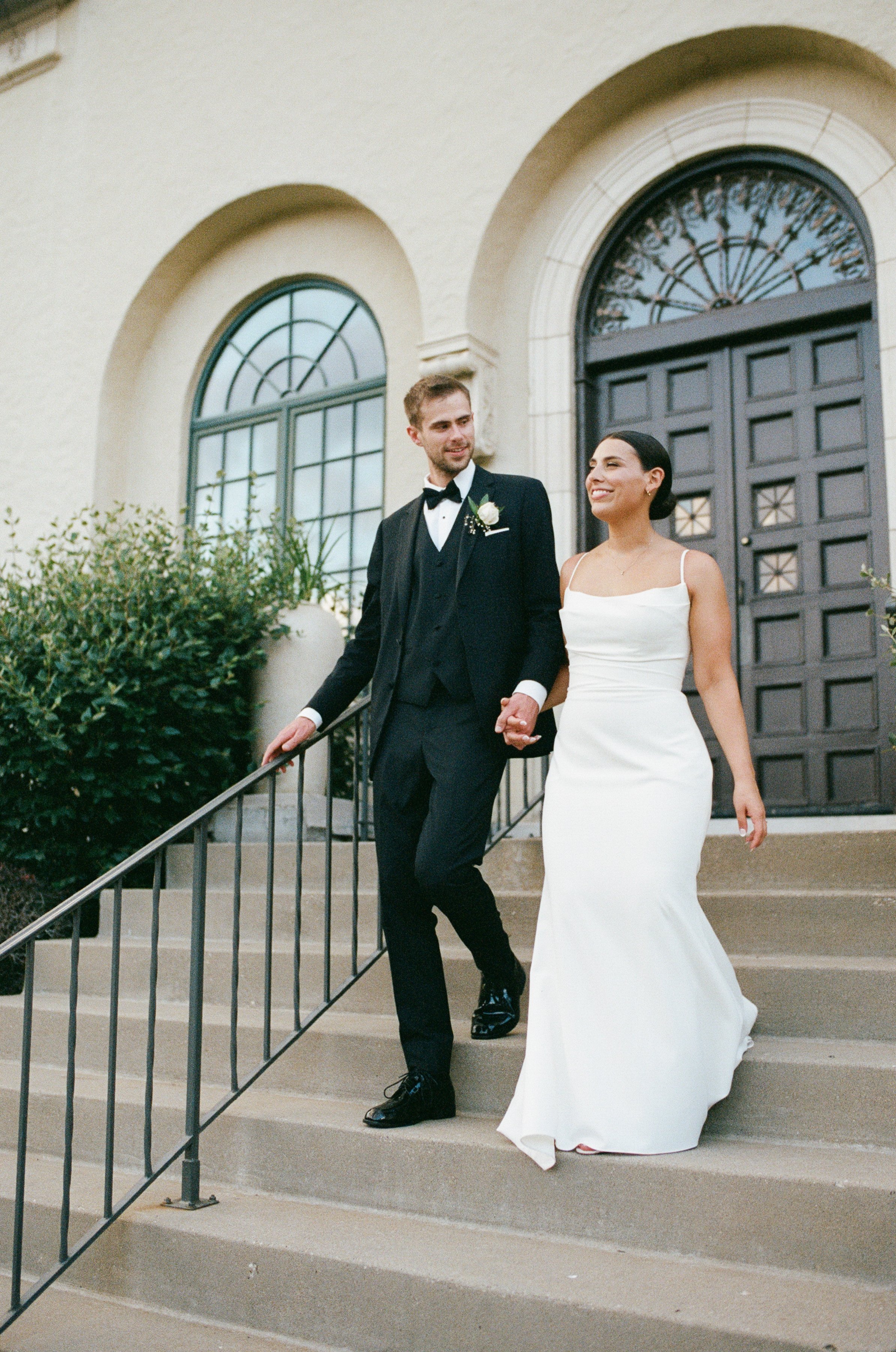 A newlywed couple walking down the steps of a building, holding hands, with the groom in a tuxedo and the bride in a white wedding dress.