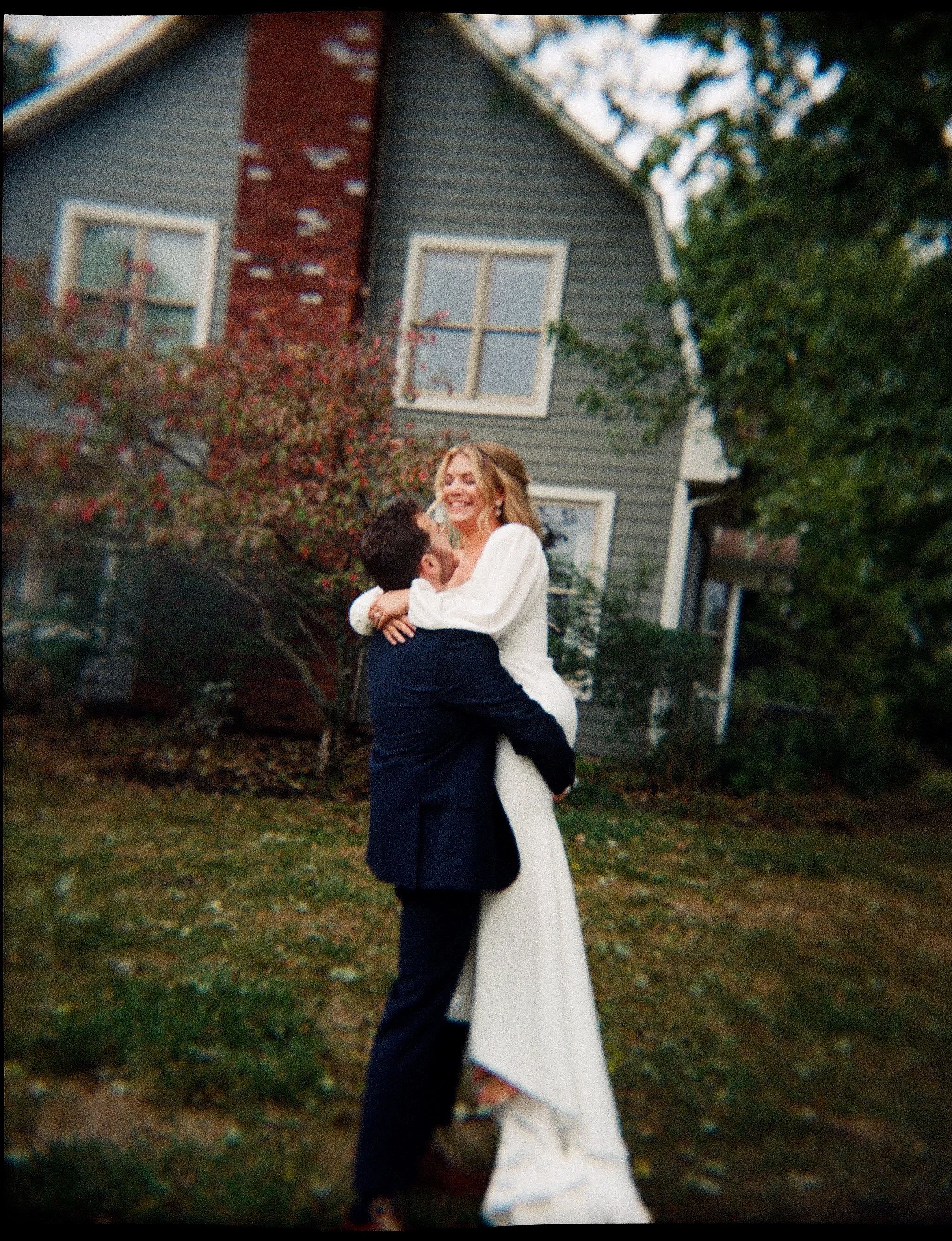 A couple in wedding attire embracing and smiling outside in front of a house with gray siding and a brick chimney, surrounded by trees and grass.