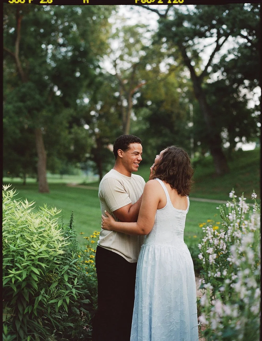 A couple in casual attire sharing an intimate moment, smiling and embracing in a lush green park with trees and flowering plants.