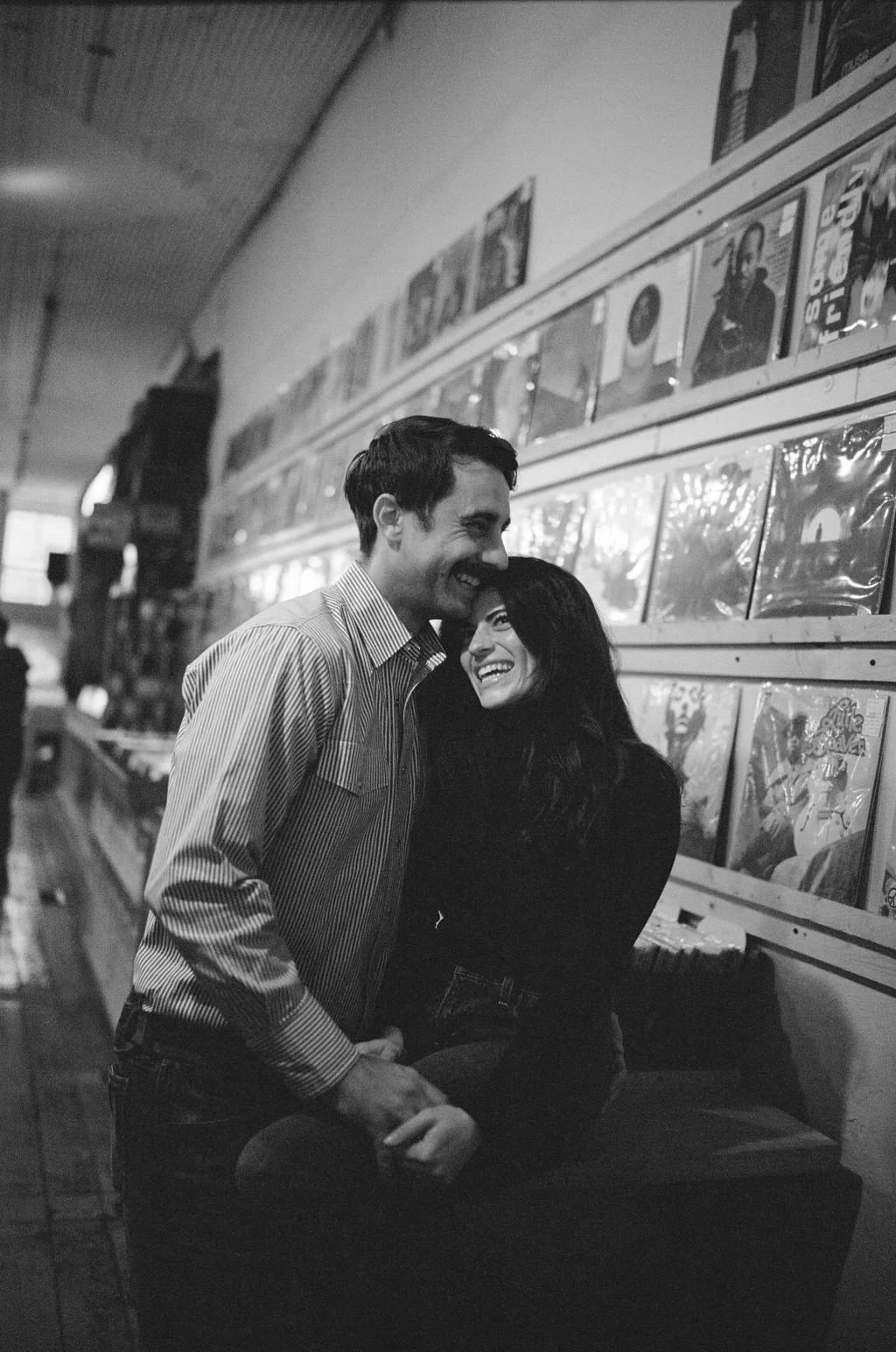 A black and white photo of a smiling couple embracing each other in a record store with shelves of vinyl records in the background.