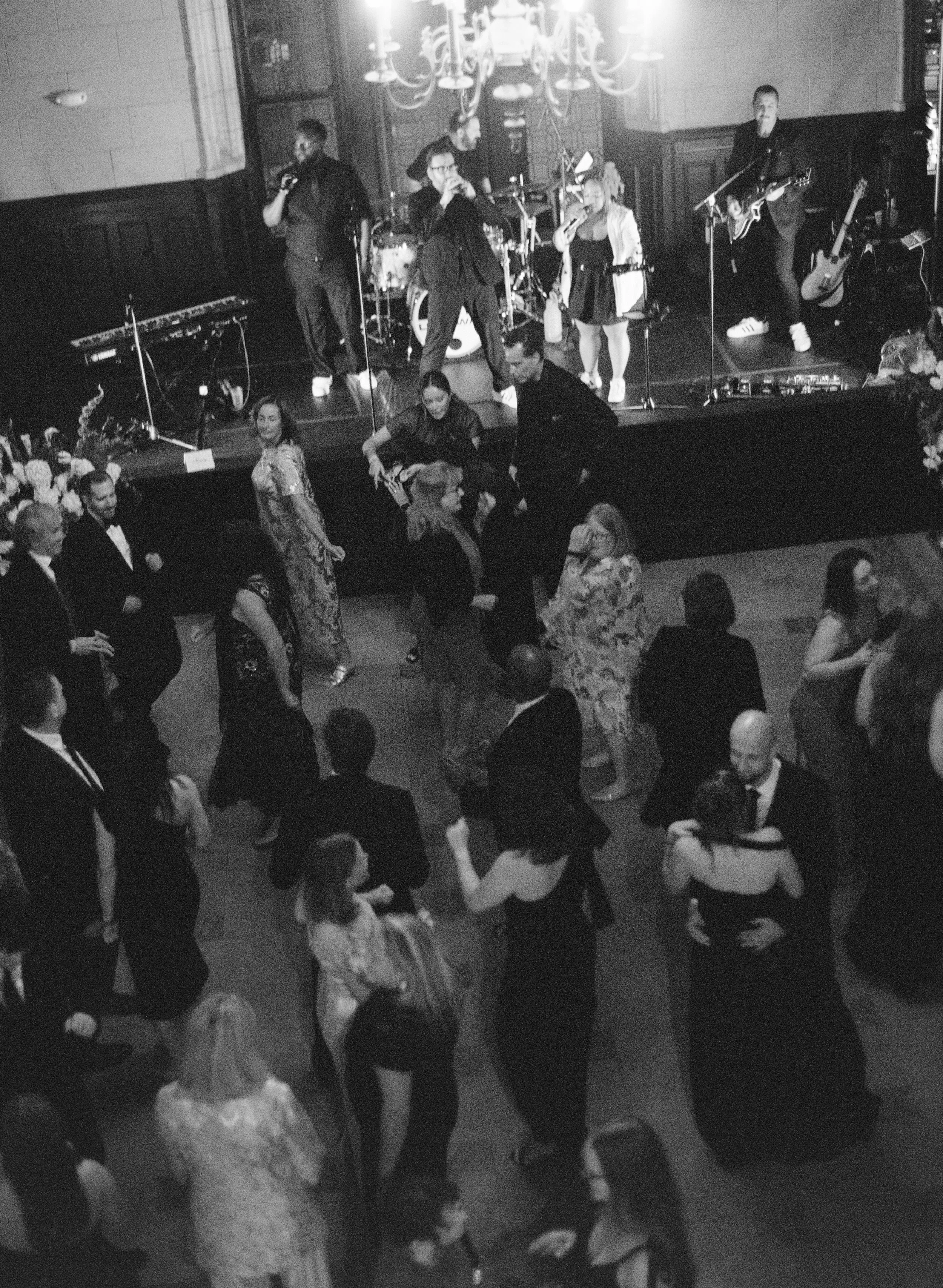 A black and white photo of people dancing on a floor in front of a stage with a band performing. The band has a drum set, keyboard, guitars, and singers, with a chandelier hanging above.