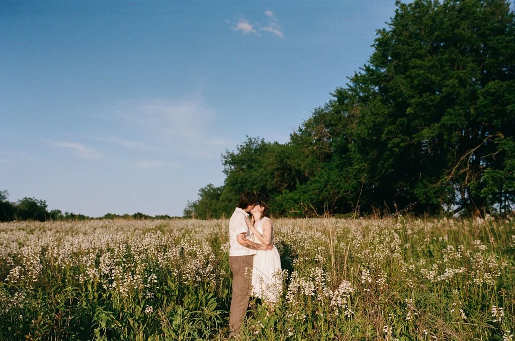 A couple embraces in a field of wildflowers with tall grass, trees, and a clear blue sky in the background.