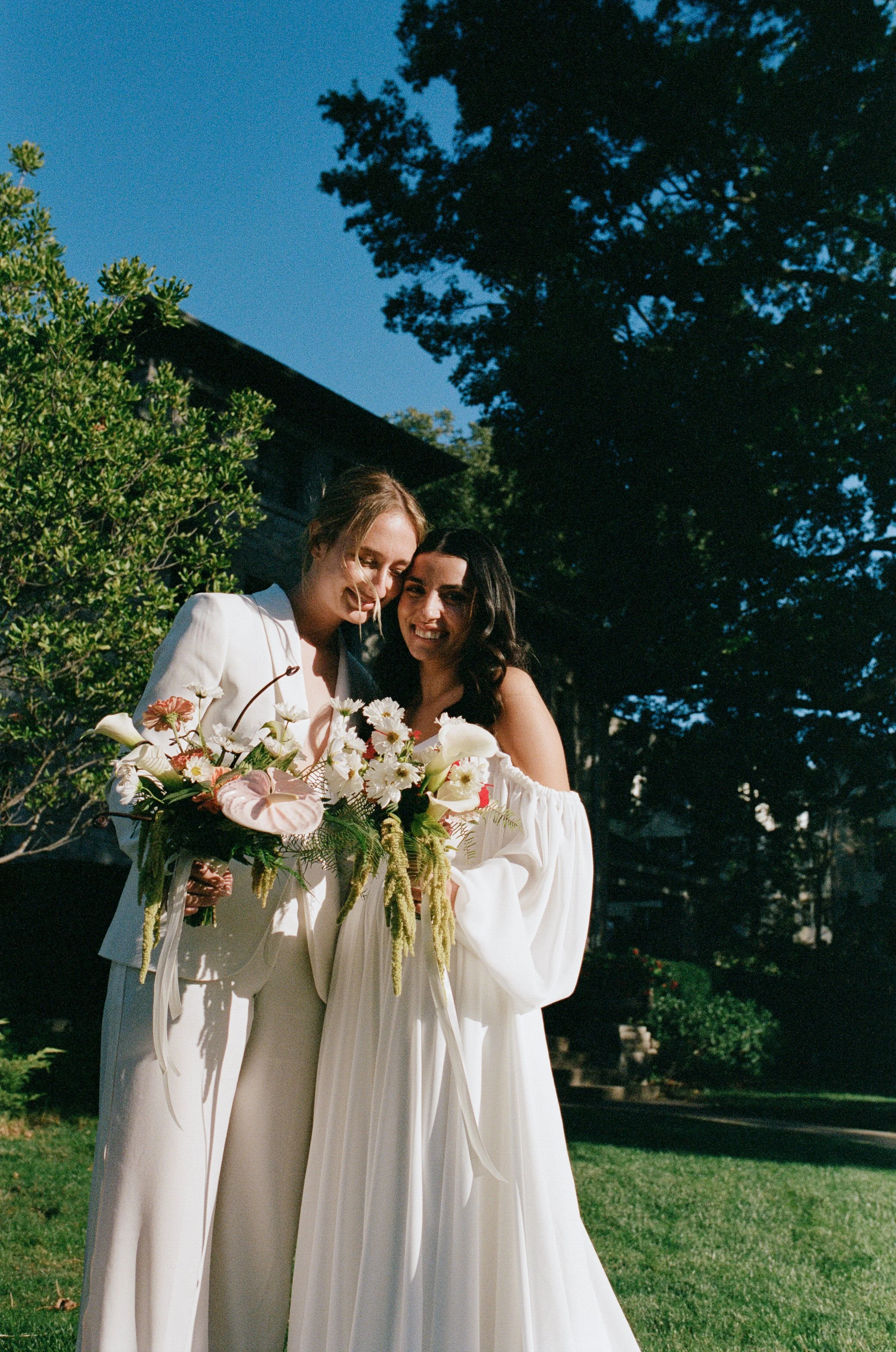 Two women in wedding attire, smiling and hugging outdoors, holding a bouquet of flowers against a backdrop of trees and a clear blue sky.