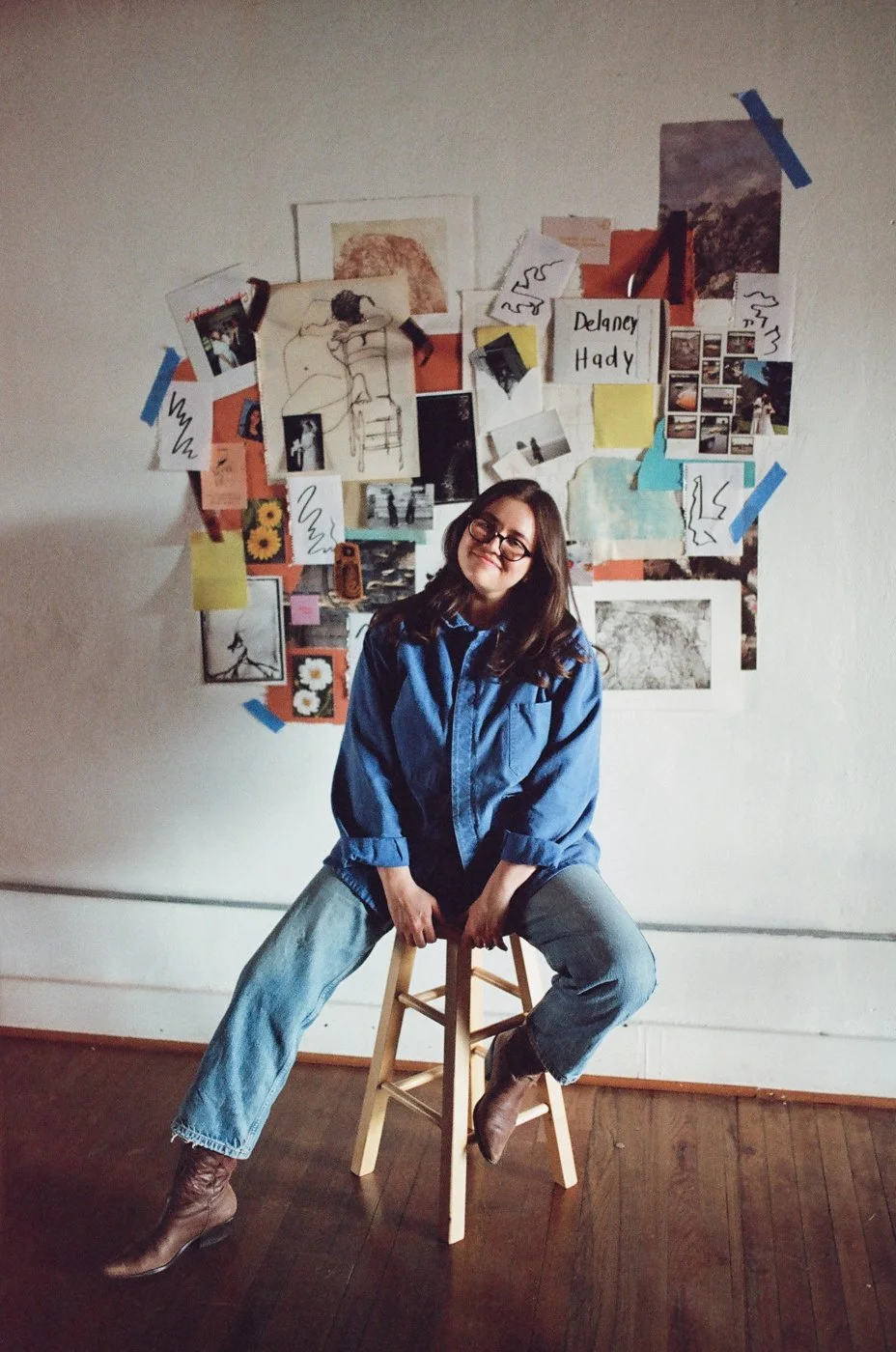 A woman with long brown hair and glasses, wearing a blue jacket and jeans, sits on a wooden stool in front of a collage on a white wall. The collage includes photos, drawings, and various pieces of paper, some attached with tape.