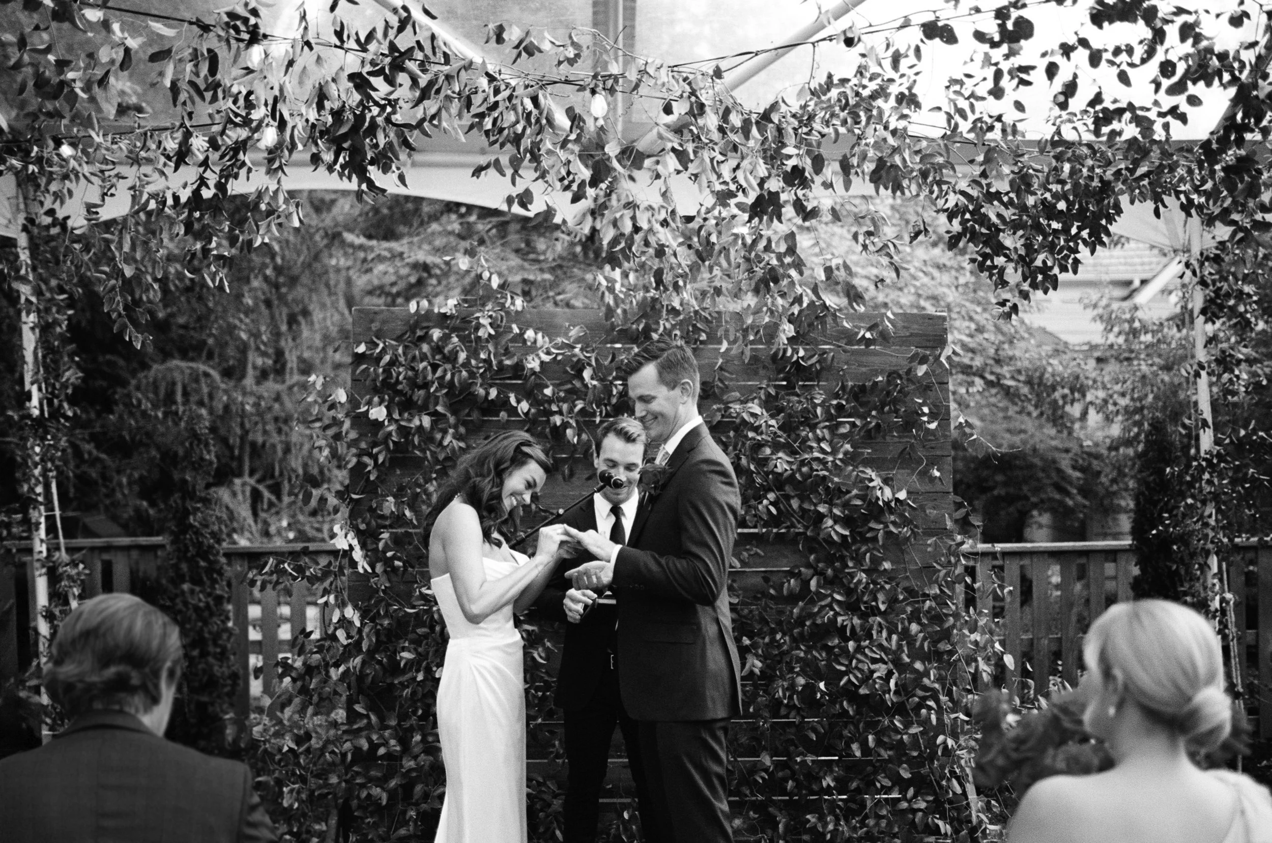 A black-and-white photo of a wedding ceremony with a bride and groom exchanging rings under a decorated arch outside, with seated guests watching.