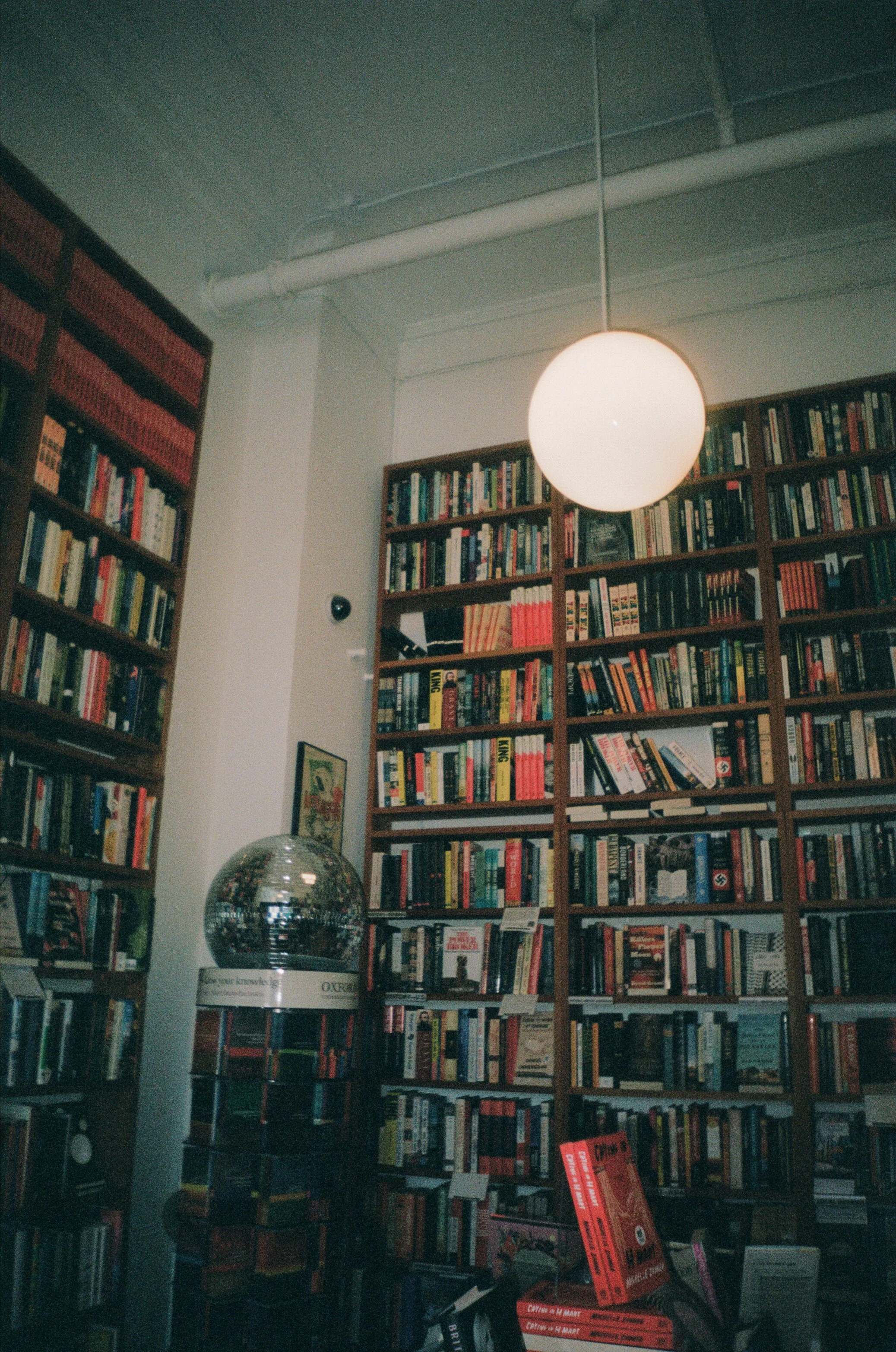Bookshelves filled with books, a round overhead light, a small framed picture, a MCM light fixture, and a red sign on display in a bookstore or library.
