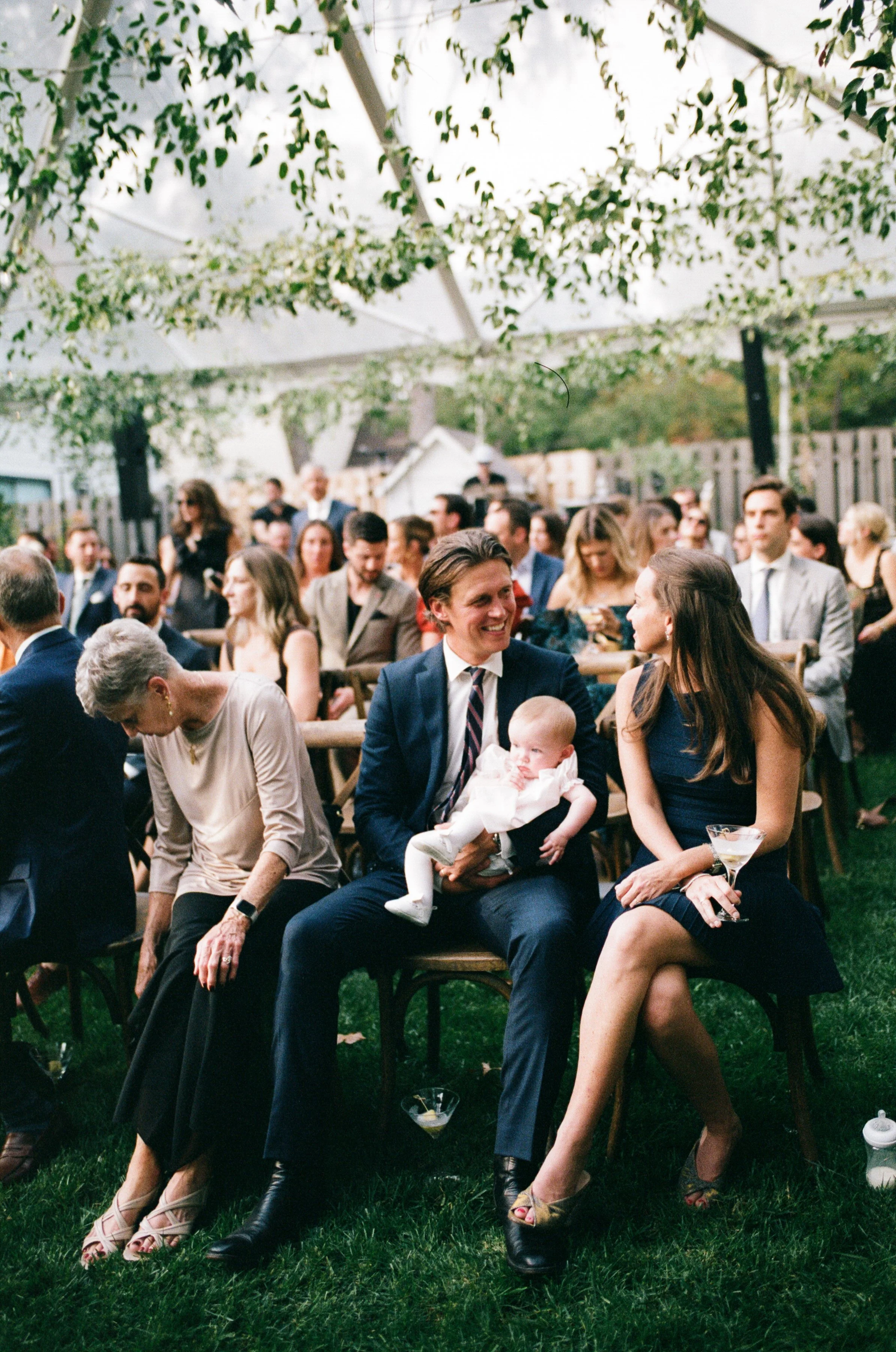 People attending an outdoor wedding or celebration under a tent with floral decorations, sitting and socializing while dressed in formal attire.