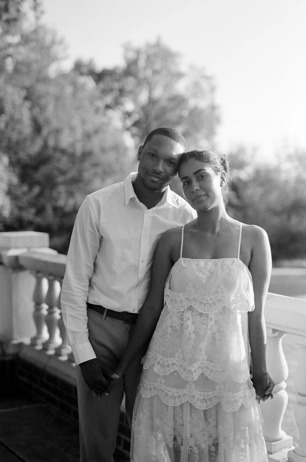 A black and white photo of a couple standing close together outdoors, holding hands, with trees in the background. A man is wearing a white shirt and light-colored pants, and a woman is wearing a light, lace dress.