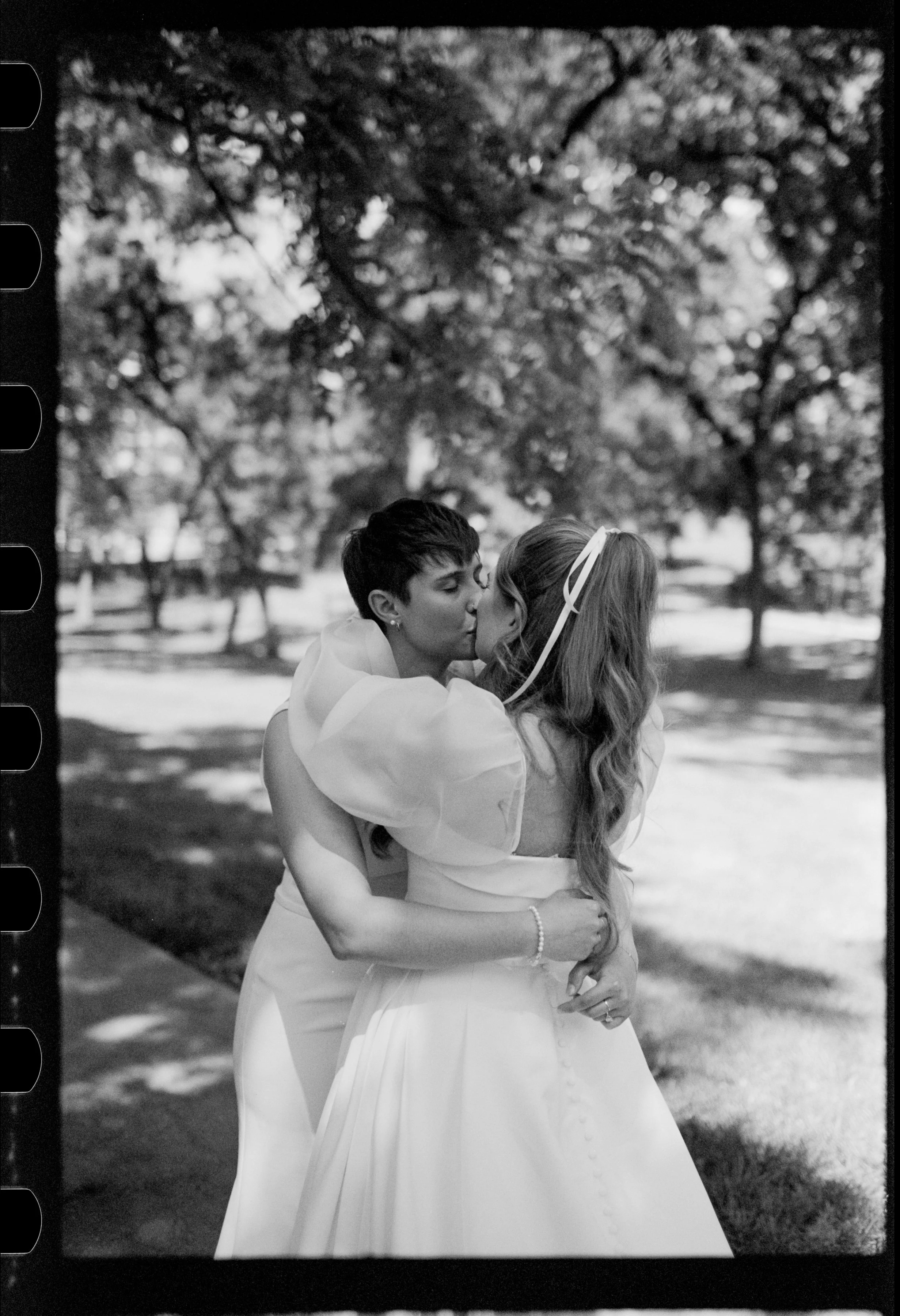 Two women, one with short dark hair and the other with long wavy hair, are kissing outdoors on a sunny day. They are dressed in wedding gowns, embracing in a park with trees in the background.