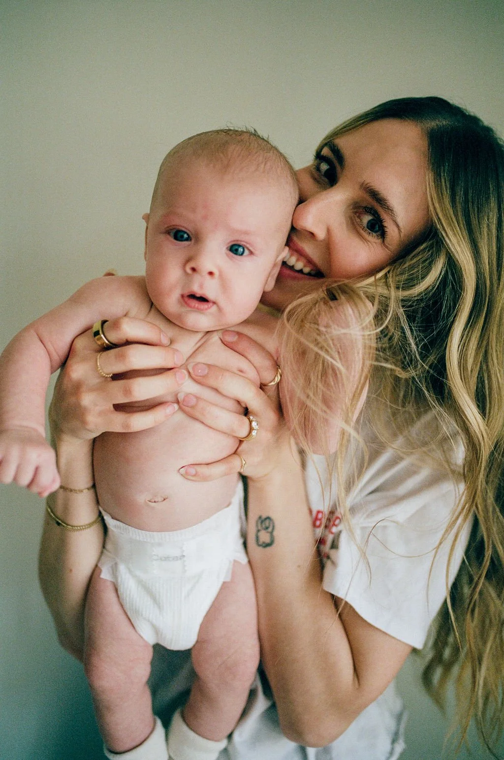 A woman with long wavy blonde hair holding a baby with short hair and blue eyes, both smiling. The woman is wearing rings and has a small tattoo on her arm. The baby is in a diaper.