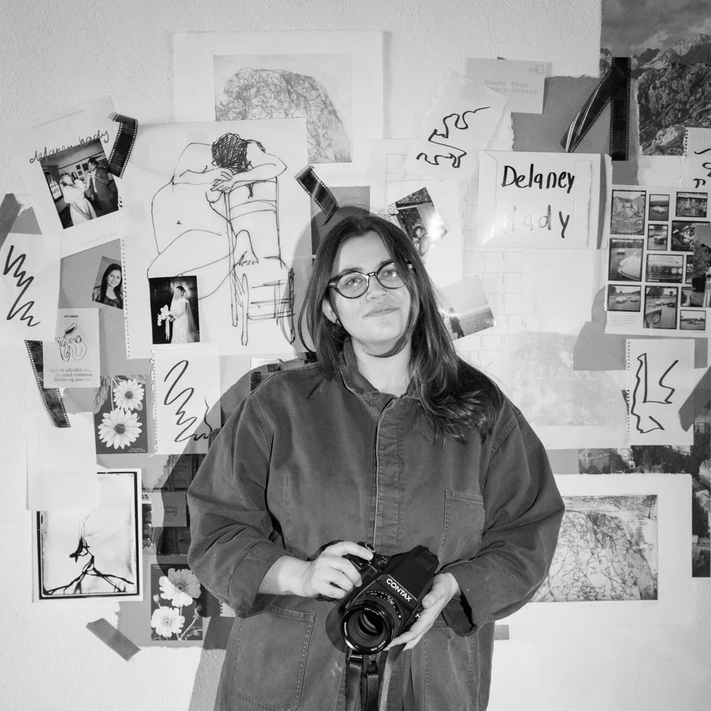 Delaney Hady holding a camera stands in front of a collage wall with drawings, photographs, and notes.