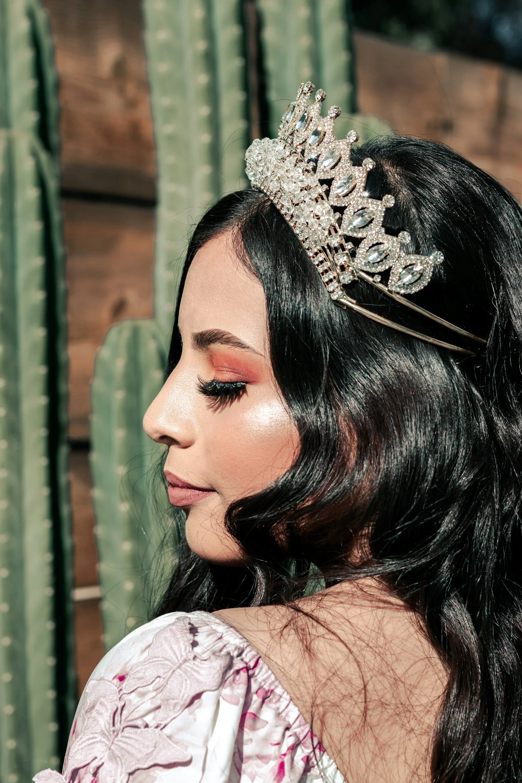 A woman with long black hair wearing a silver tiara. She has makeup with pink eyeshadow, long eyelashes, and a shimmery complexion. She is dressed in a white embroidered dress, back view. Background of green cacti and wooden wall.