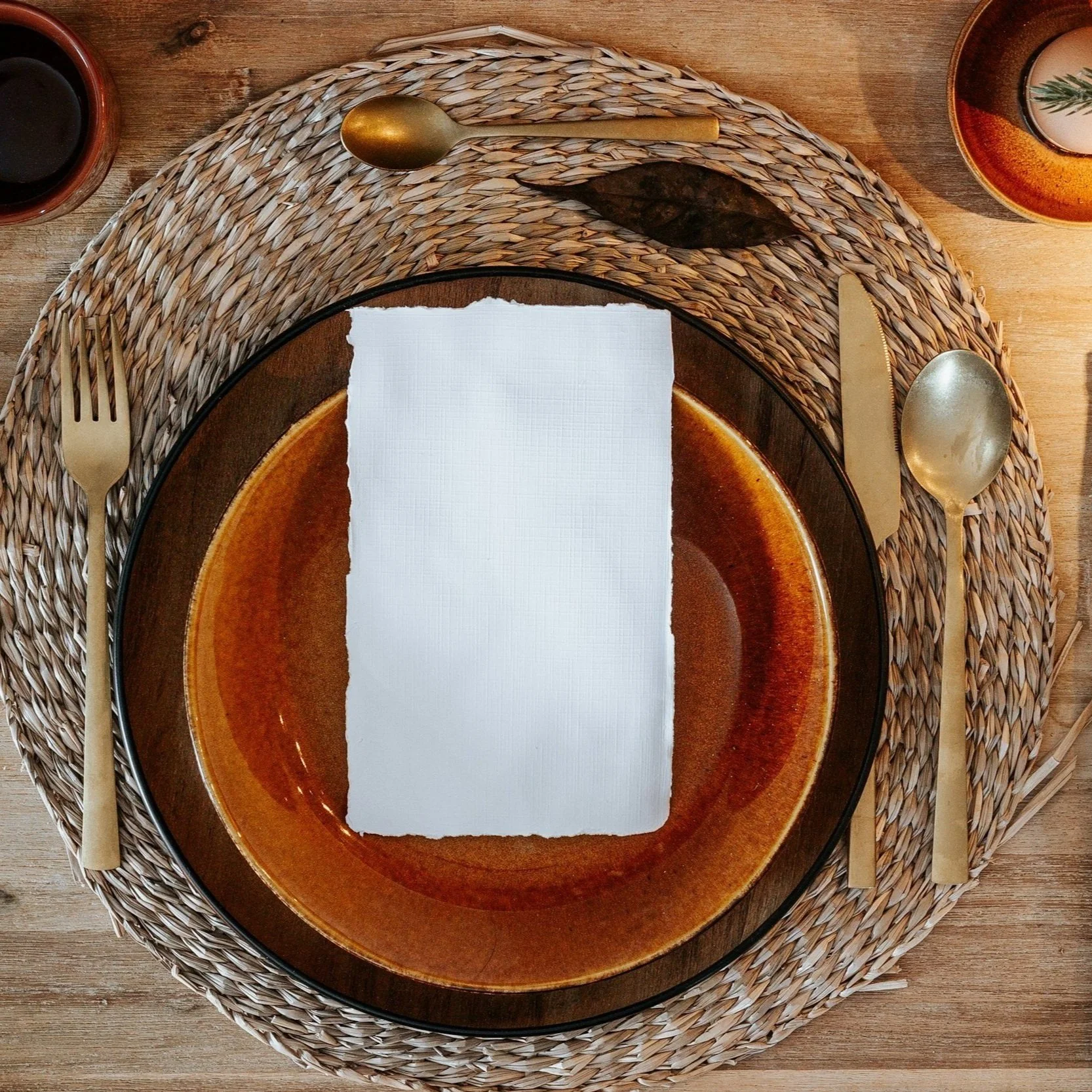 A place setting with a large round orange ceramic plate, a torn white paper napkin on top, surrounded by gold-colored cutlery on a woven placemat, on a wooden table.
