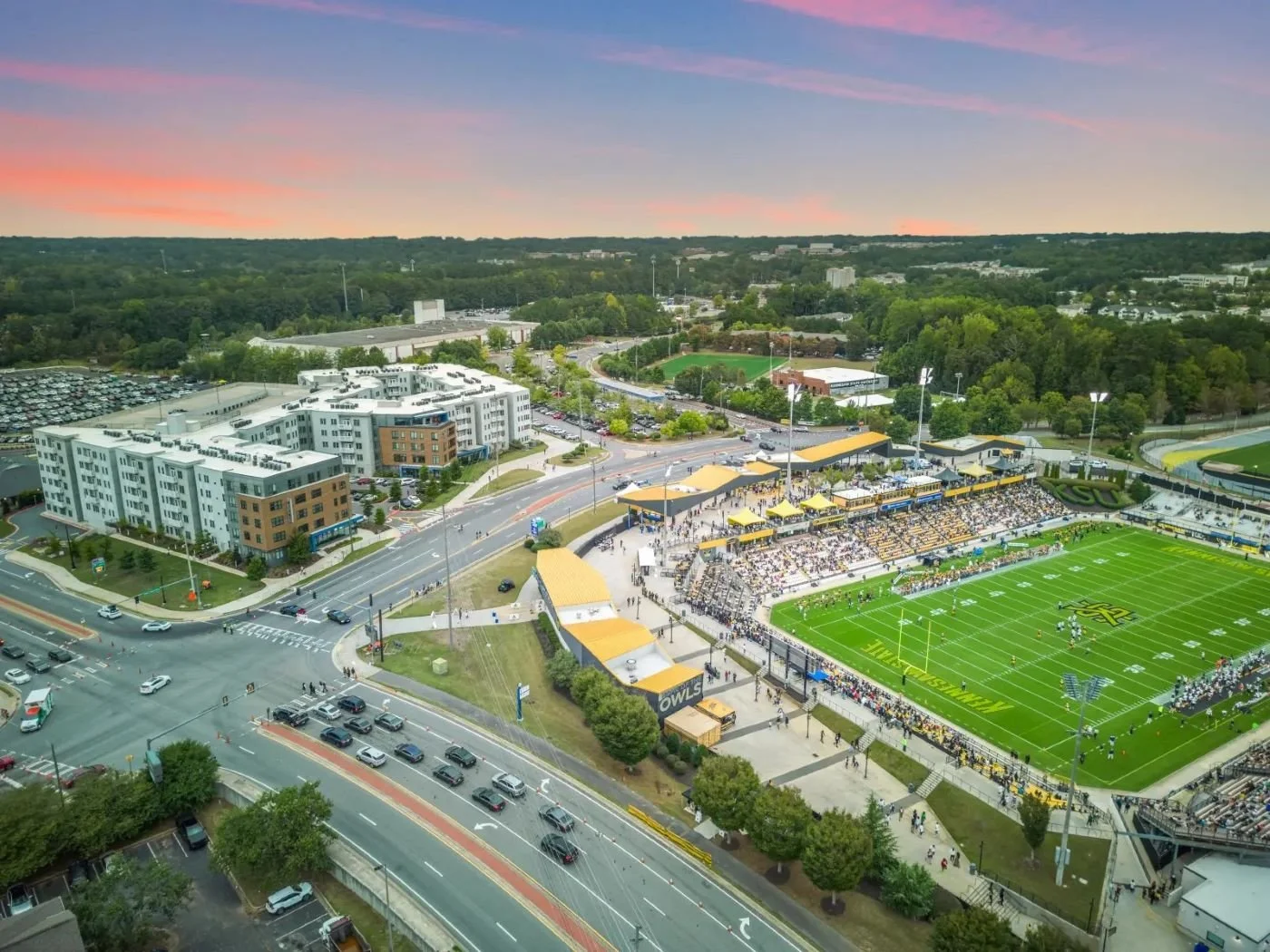 Aerial view of a football stadium filled with spectators during a game at sunset, surrounded by roads, a modern residential building, and green parks.