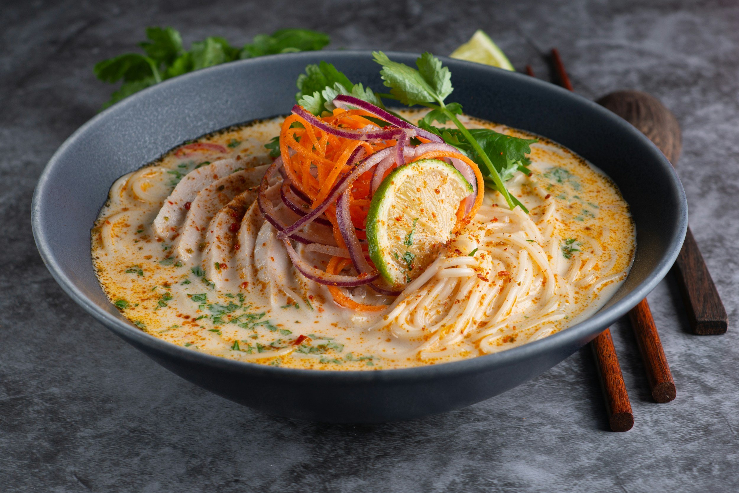 Bowl of coconut curry soup with noodles, sliced chicken, lime, cilantro, red onion, and carrot garnish.