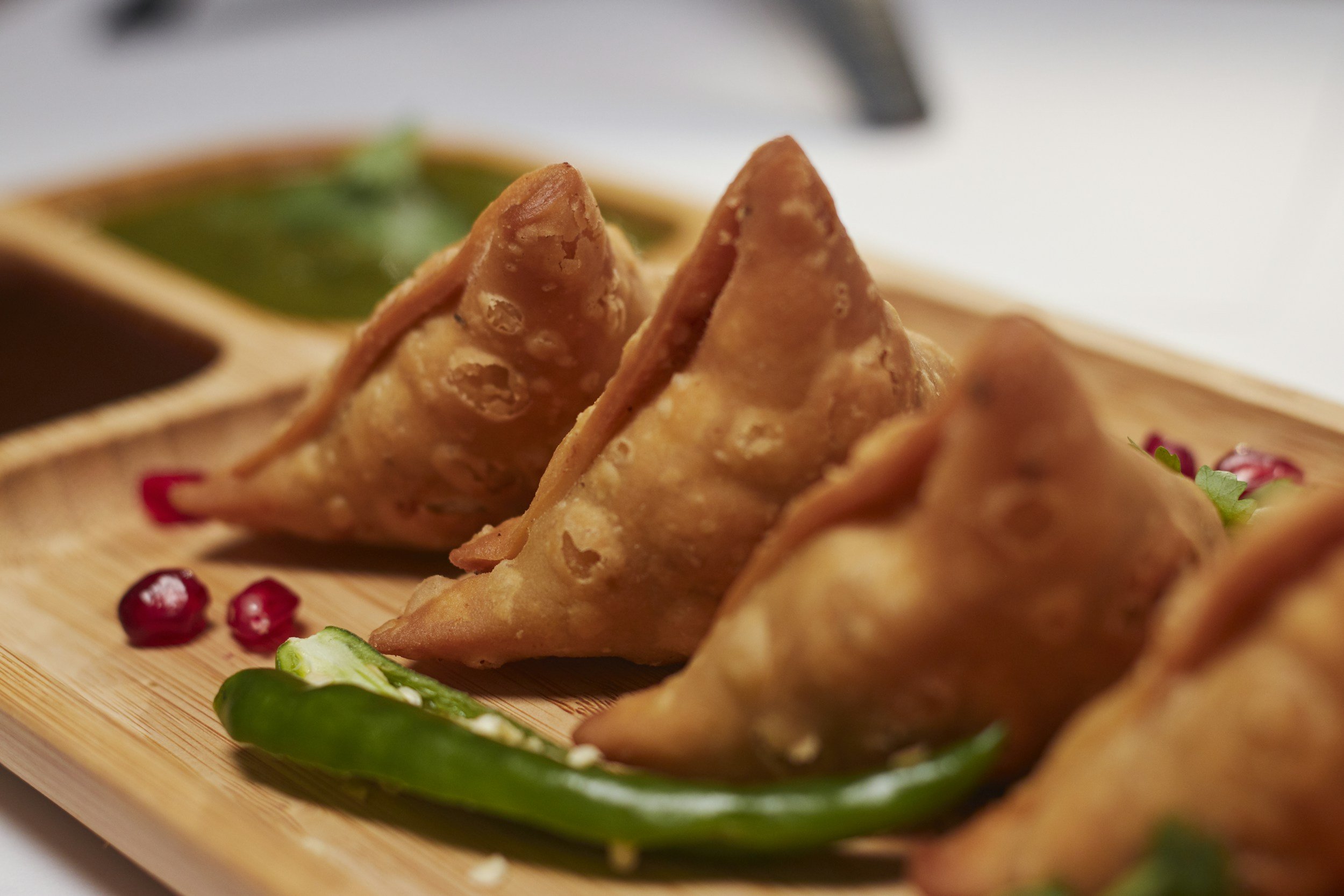 Close-up of fried samosas on a wooden plate with green chilies and pomegranate seeds, accompanied by dipping sauces in the background.