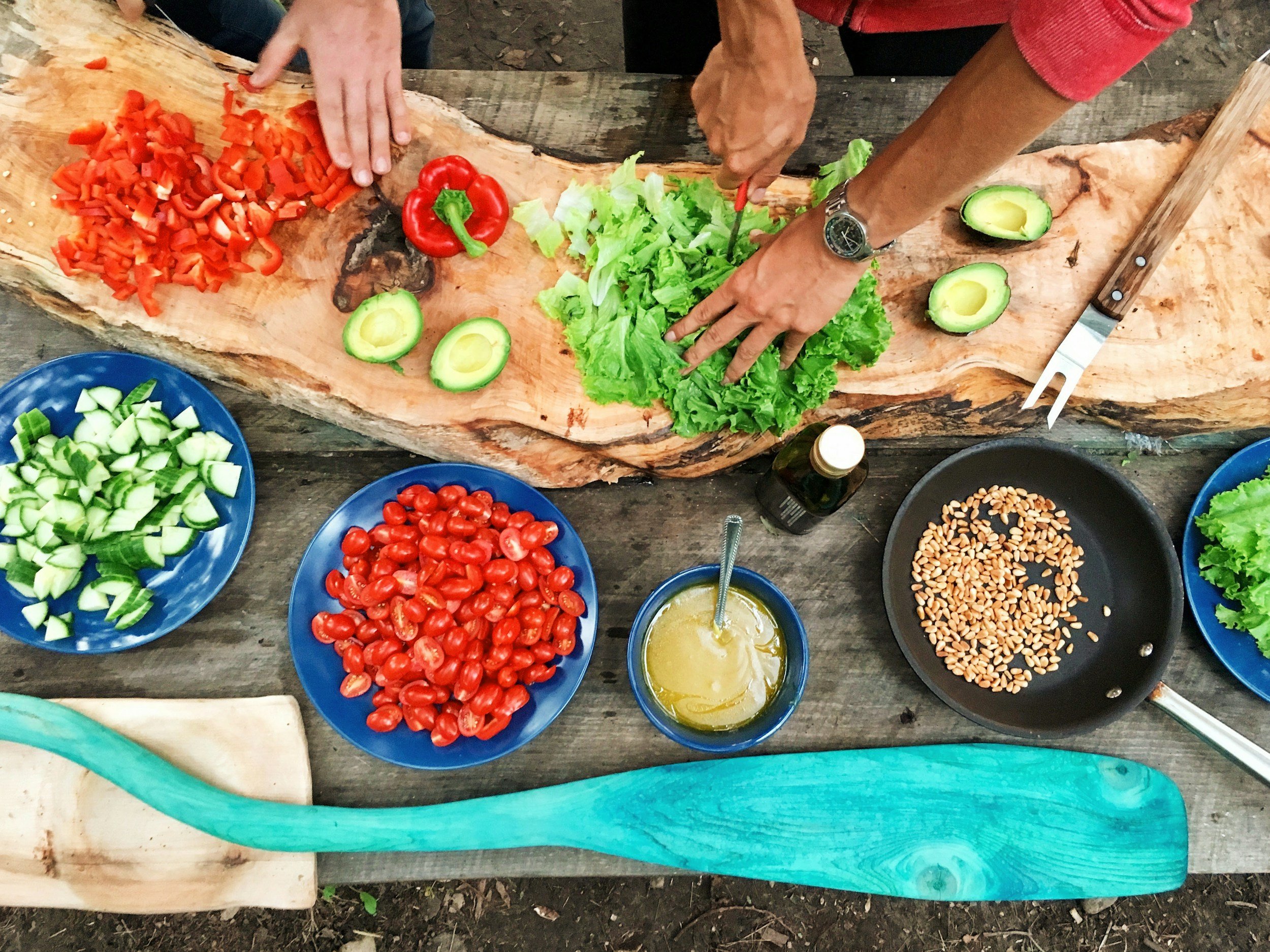 People preparing salad ingredients on a wooden surface, including chopped red bell peppers, avocado, lettuce, cucumbers, cherry tomatoes, pine nuts in a pan, with a bottle of olive oil and a bowl of dressing.