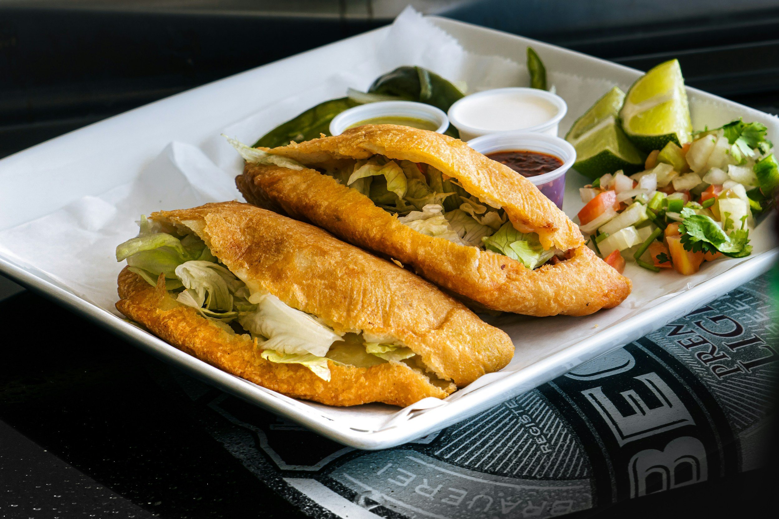 Plate of two pambazos stuffed with lettuce on a white dish, accompanied by lime wedges, salsa in small containers, and diced vegetable garnish.