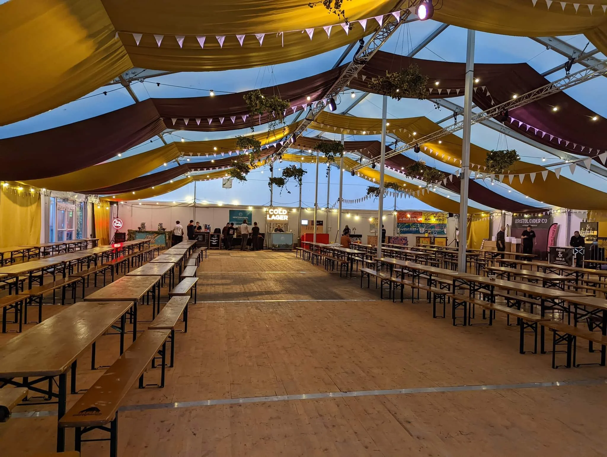 Interior view of a large festival tent with colorful fabric drapes and string lights. Long wooden tables and benches are arranged on the wooden floor, with a few people standing near the bar area in the background.