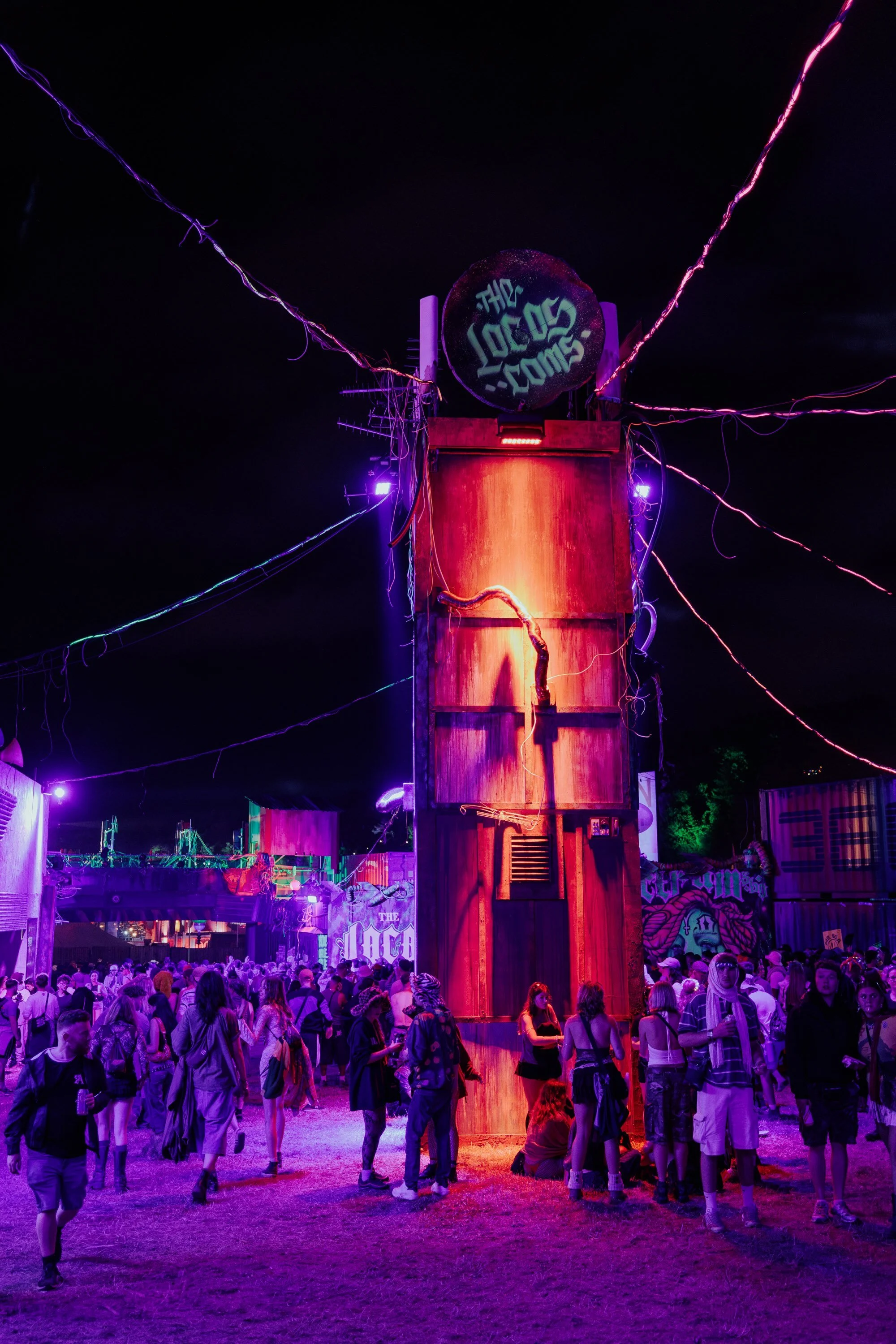 People attending an outdoor music festival at night, illuminated by colorful purple and pink stage lights, gathered around a tall, rusty, rectangular structure with wiring and a sign at the top that reads 'The Locust.'