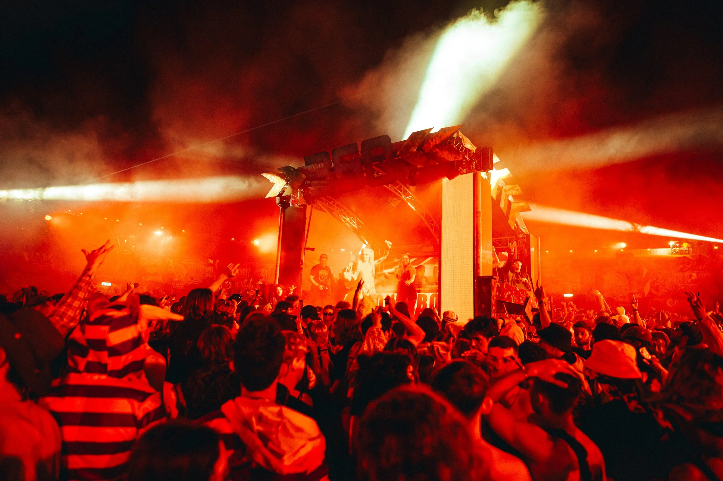 A crowded outdoor music concert at night with a large stage illuminated in red and orange lighting. Smoke and bright stage lights are visible, with performers on stage and an audience dancing and raising their hands.
