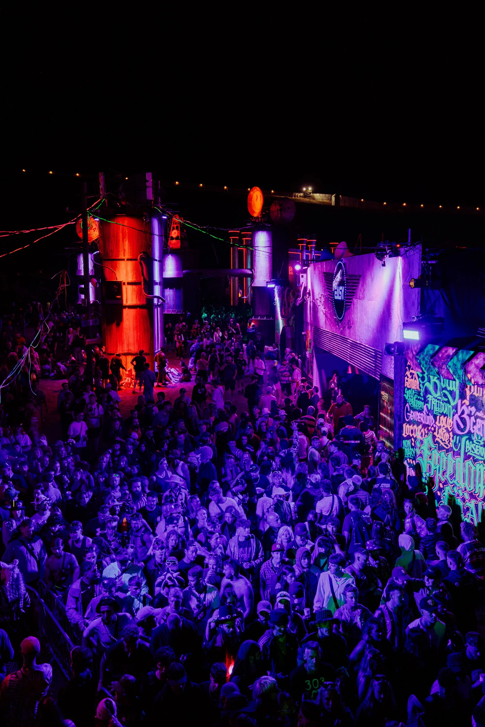 Crowd of people at a nighttime electronic music festival with colorful neon lights and graffiti on walls.