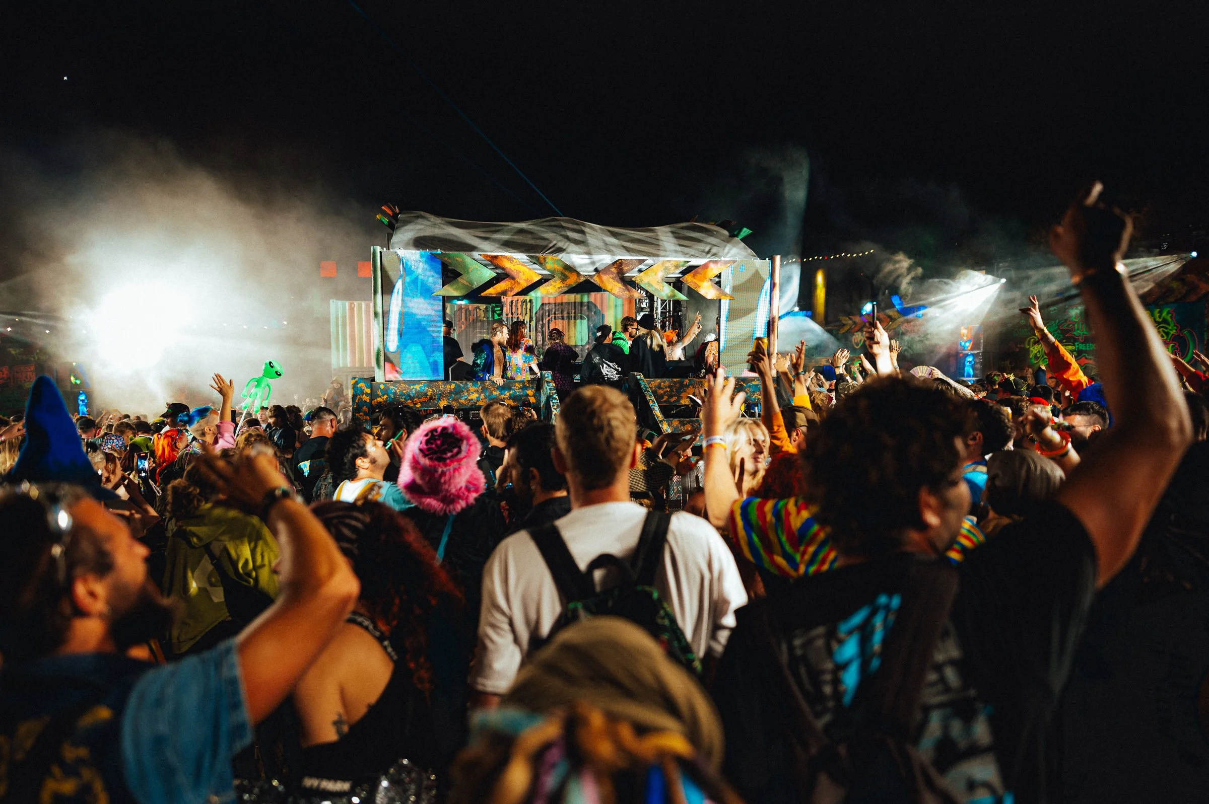 Crowd of people dancing and raising their hands at a nighttime outdoor concert or music festival with a colorful DJ stage and bright lights.