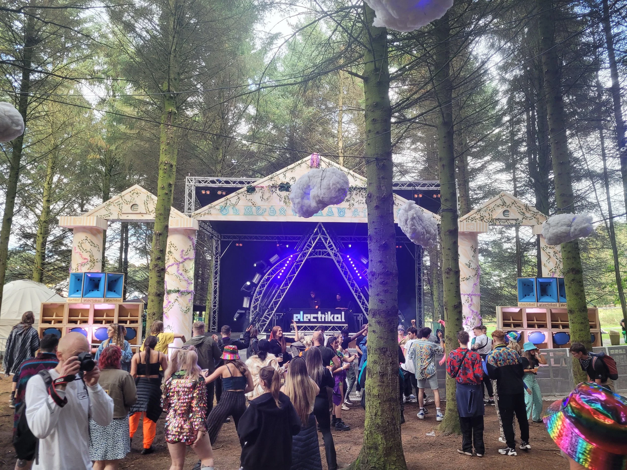 Outdoor music festival stage in a forest with people dancing and socializing, decorated with hanging cloud-like objects and large speakers.