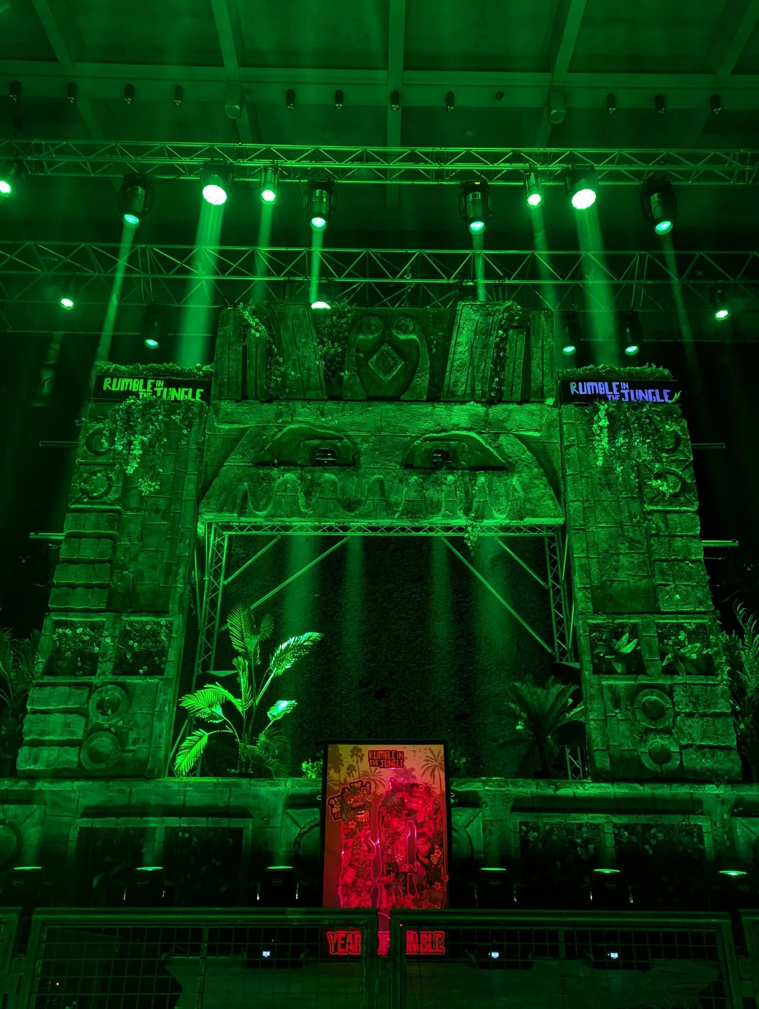 Stage setup with green lighting, featuring a tropical theme with plants and a large stone arch, for an event called 'Rumble in the Jungle'.