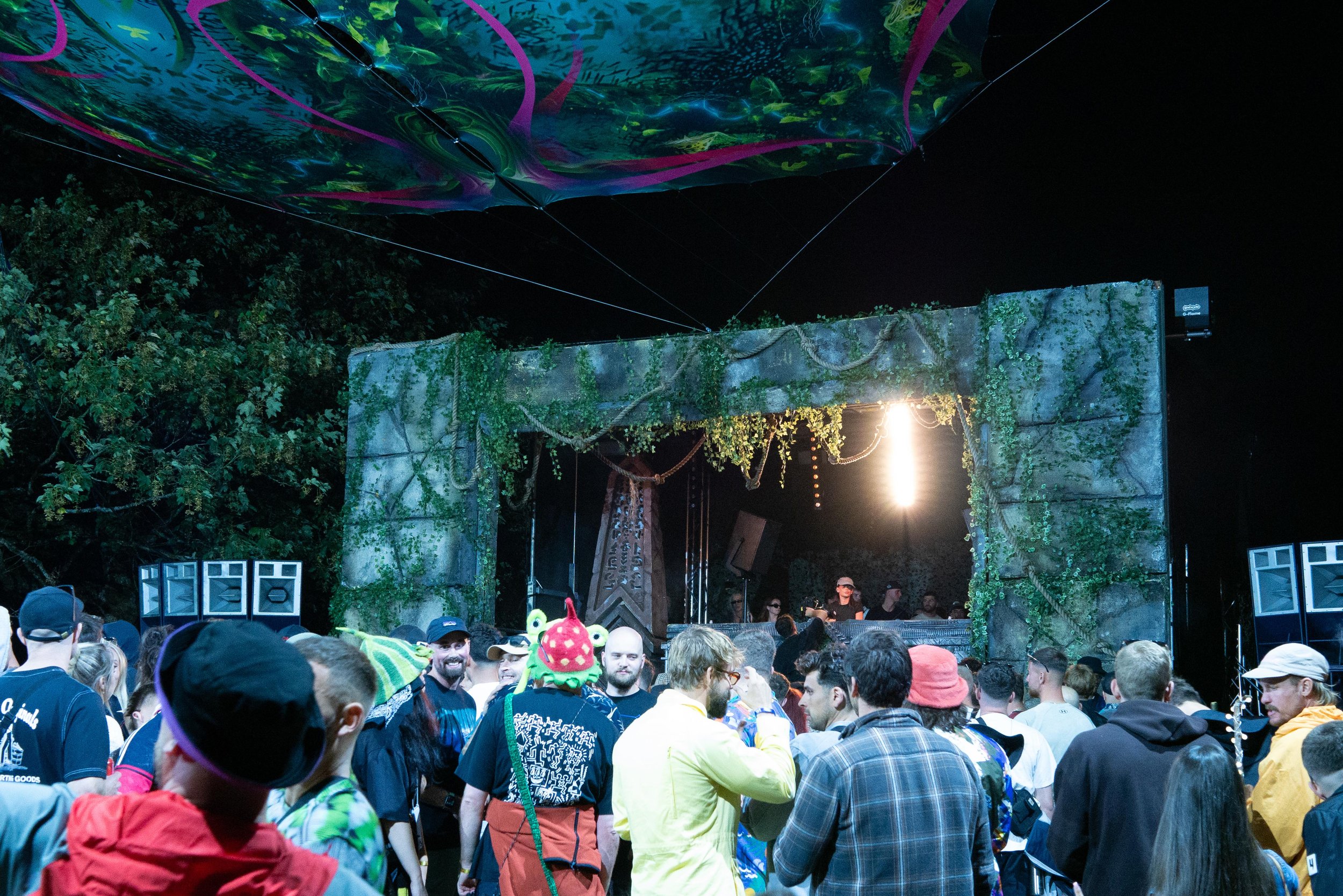 Crowd of people at an outdoor concert under a black night sky. The stage has a decorative backdrop with green foliage and vines, and colorful lighting. Some audience members are wearing hats and colorful clothing.