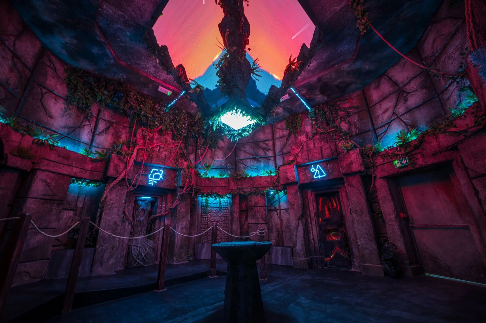 Neon-lit room with an artificial rock cave design, featuring illuminated signs, plants, and a ceiling mural of a sunset over mountains.