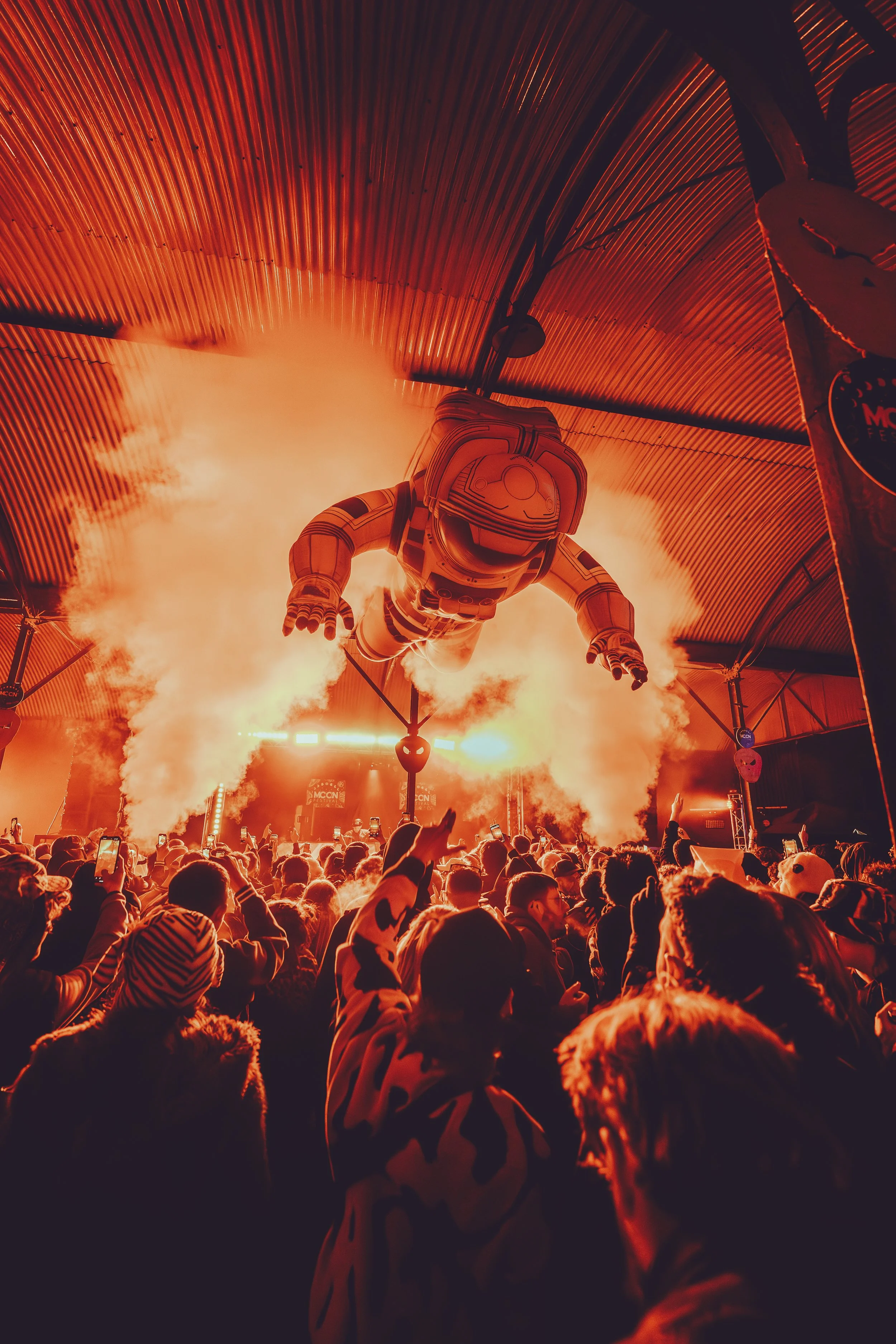 A large crowd of people at an indoor event with an inflatable astronaut figure hanging from the ceiling, illuminated by orange lighting and smoke effects.