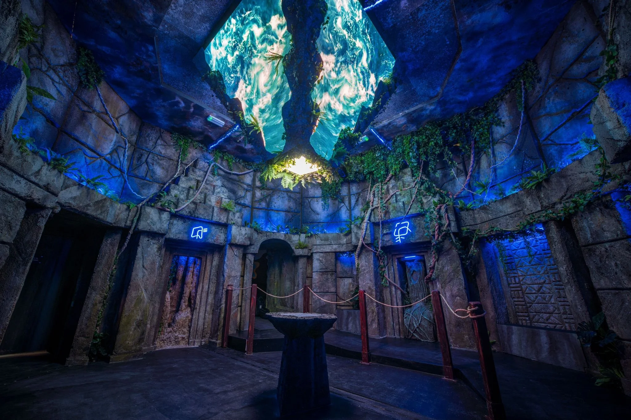 Indoor exhibit with stone walls, vines, and glowing blue signs, featuring a patch of sky and clouds projected on a ceiling