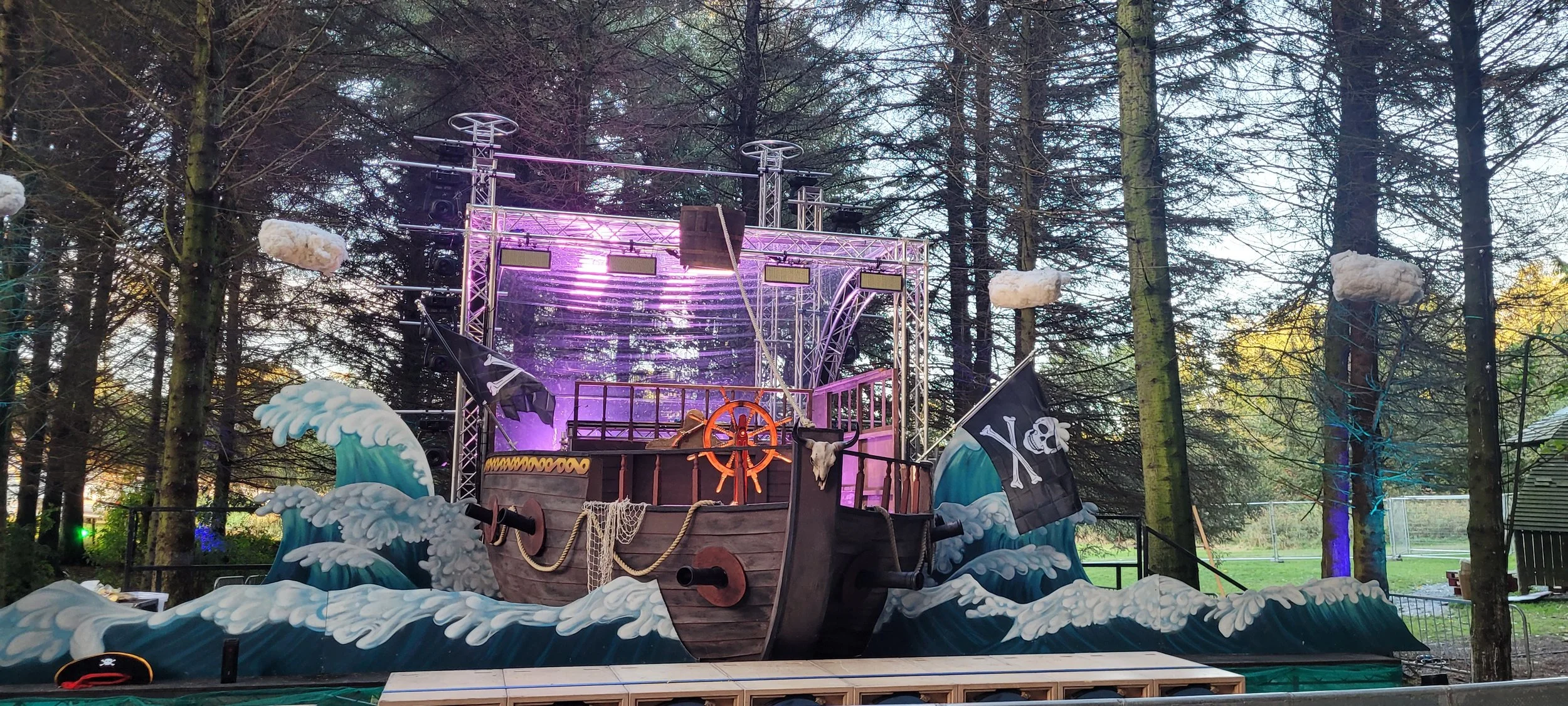 The stage is designed as a pirate ship with a wooden hull, ship wheel, and pirate flags, set on a platform with exaggerated ocean waves and clouds in a wooded outdoor environment.