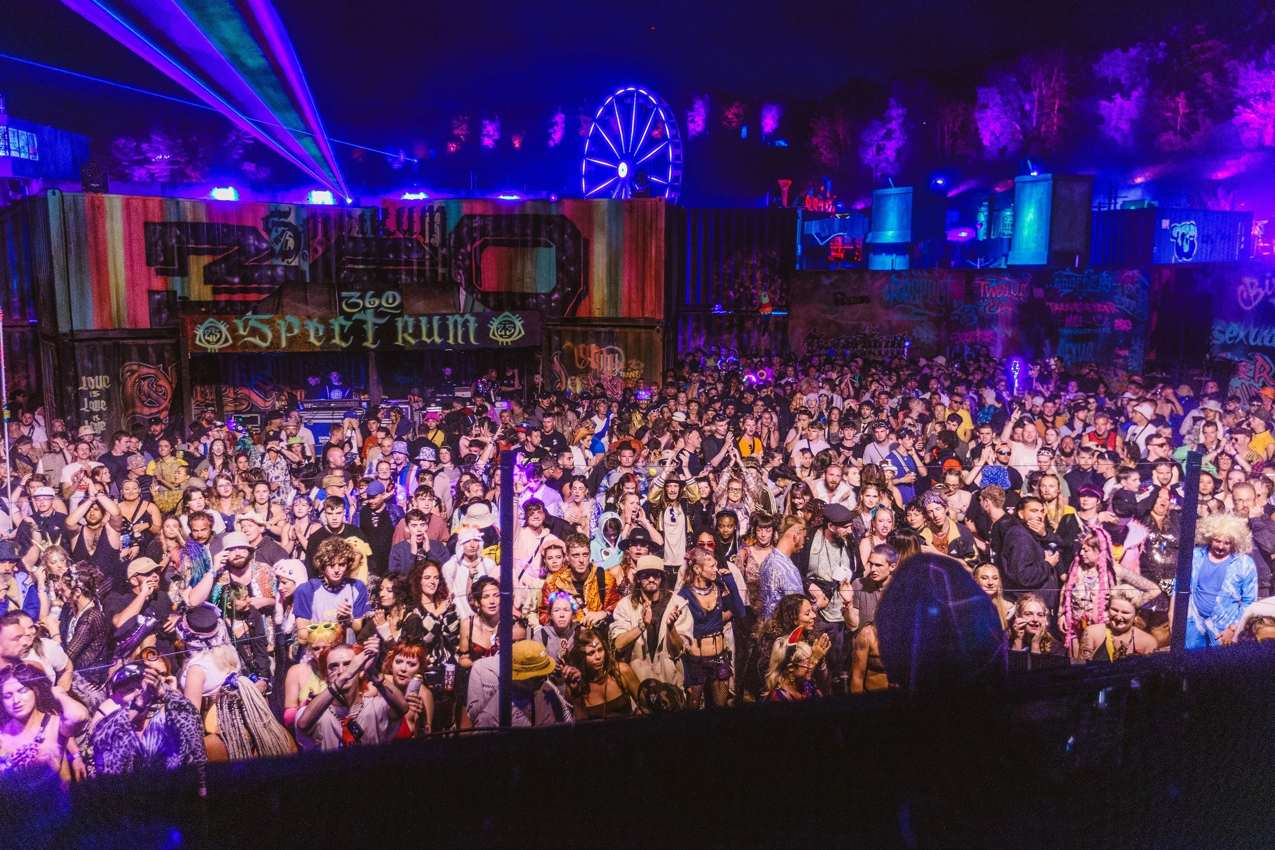 Twelve34 Productions - boomtown 2025 A large crowd at a music festival or concert, with colorful stage lighting and a Ferris wheel in the background at night.