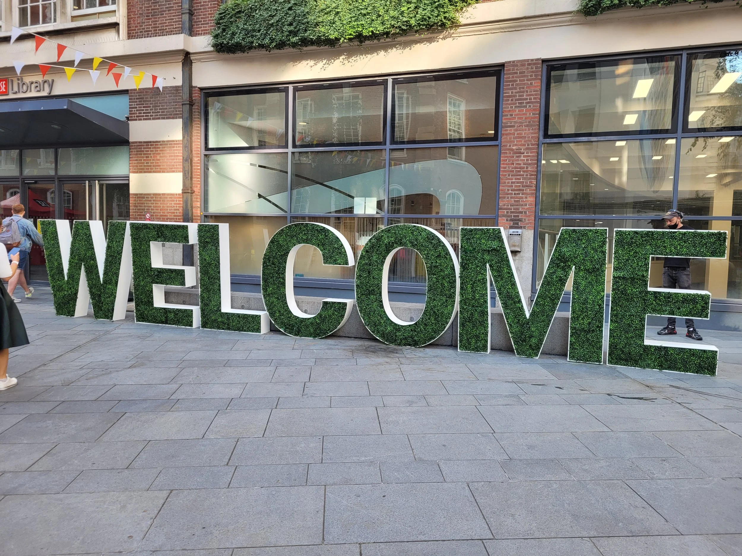 Large decorative sign spelling out 'WELCOME' with letters covered in green foliage, positioned outside a brick building with large glass windows.