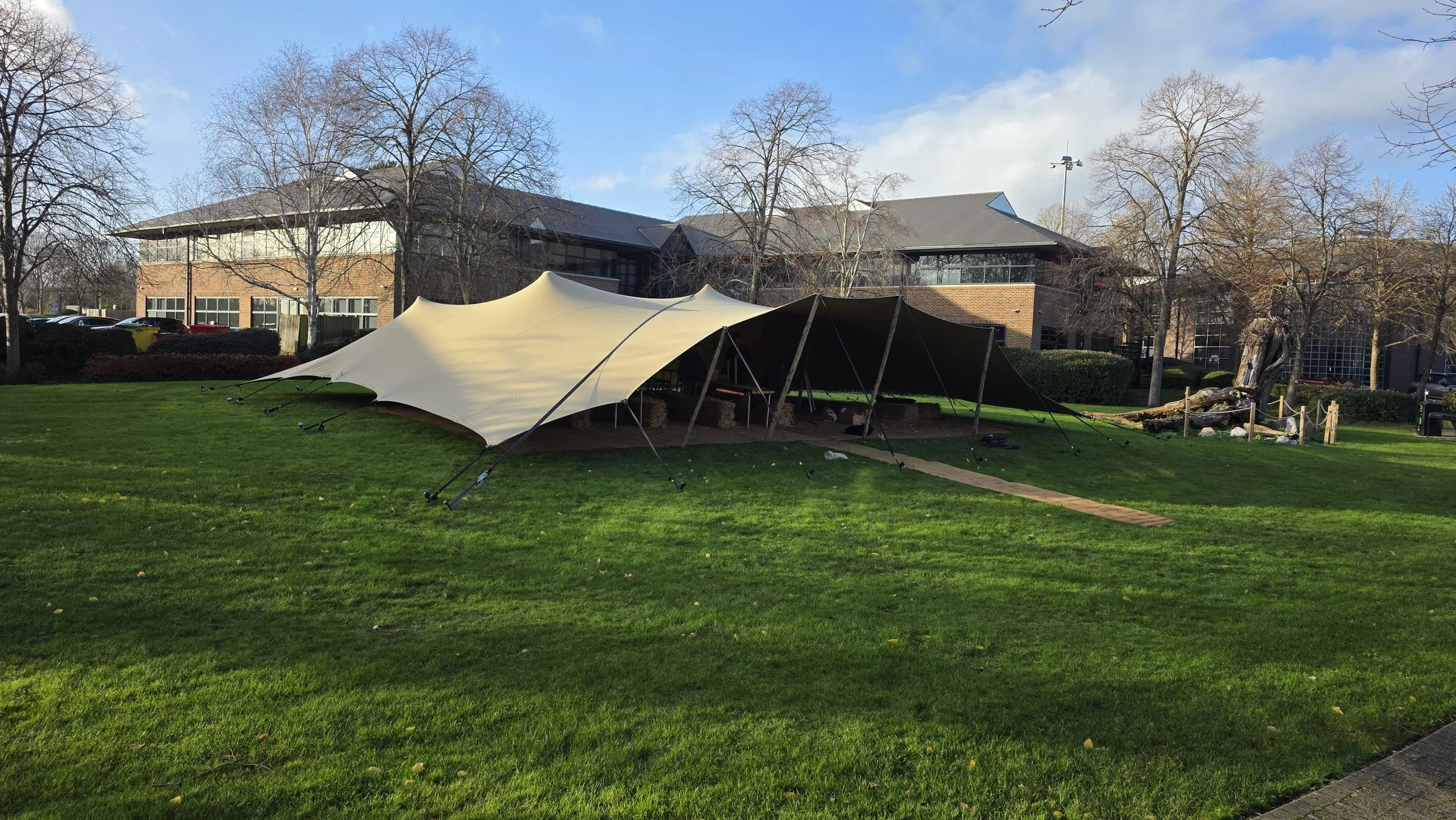 Marquee & Tent Supplier - Twelve34 Productions. A large, damaged outdoor canopy tent on a grassy lawn in front of a modern brick building with trees and blue sky in the background.