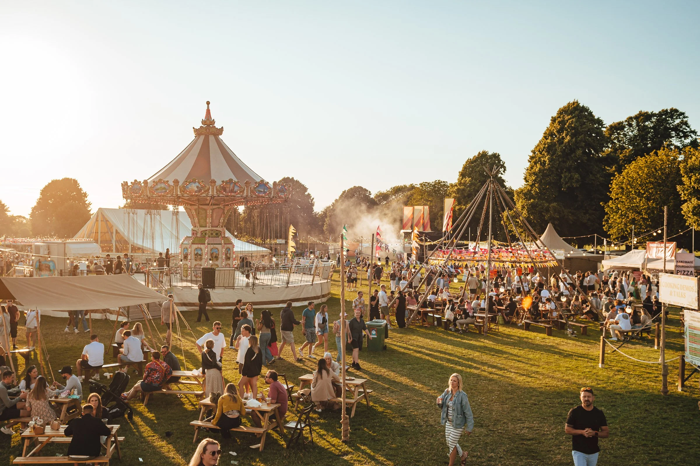 A lively outdoor fair with a vintage carousel, a swing ride, and a crowd of people enjoying food and entertainment during golden hour.