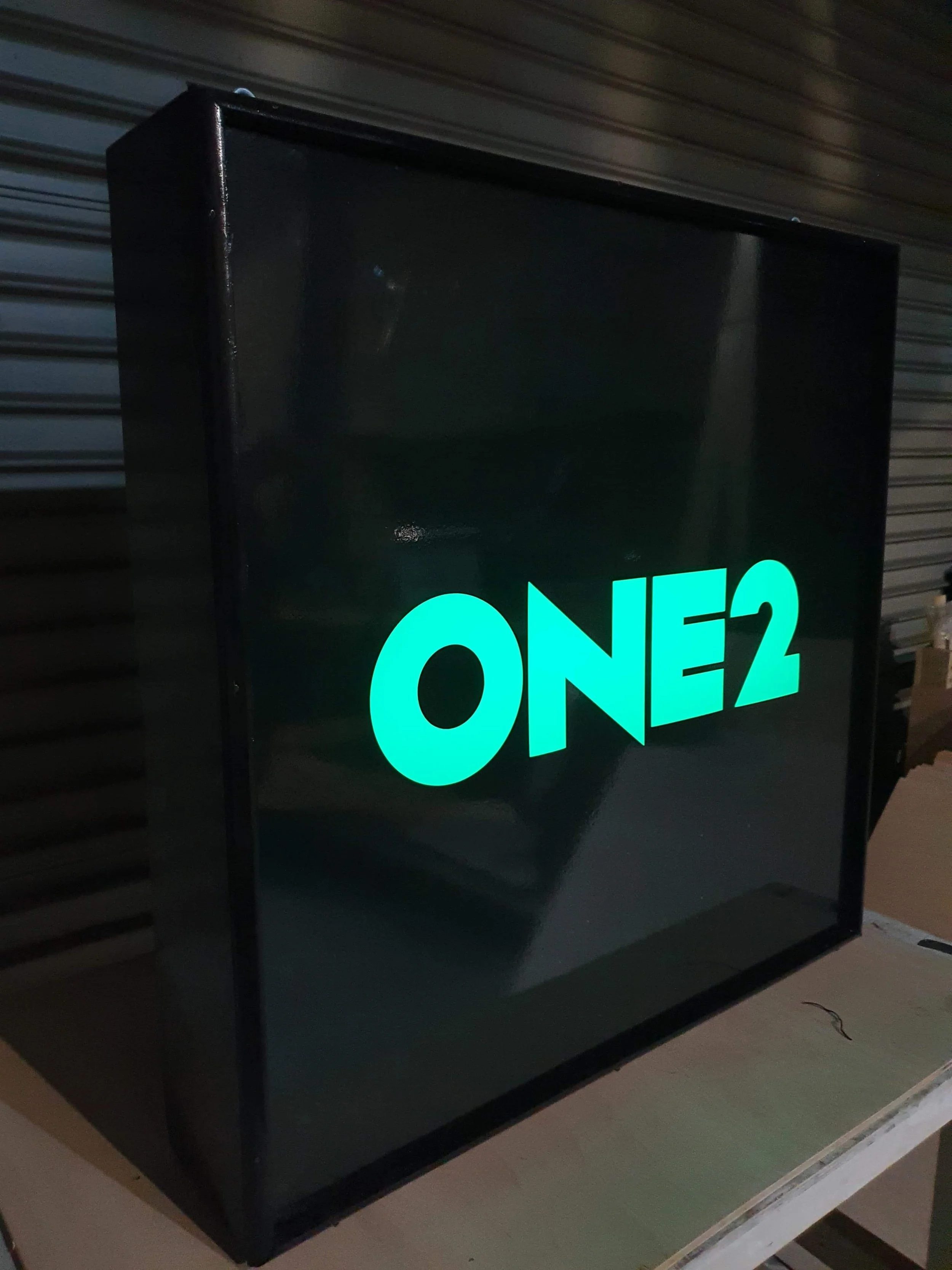  illuminated sign displaying the text 'ONE2' in bright teal letters, with a black background, placed on a table in an indoor or storage area.