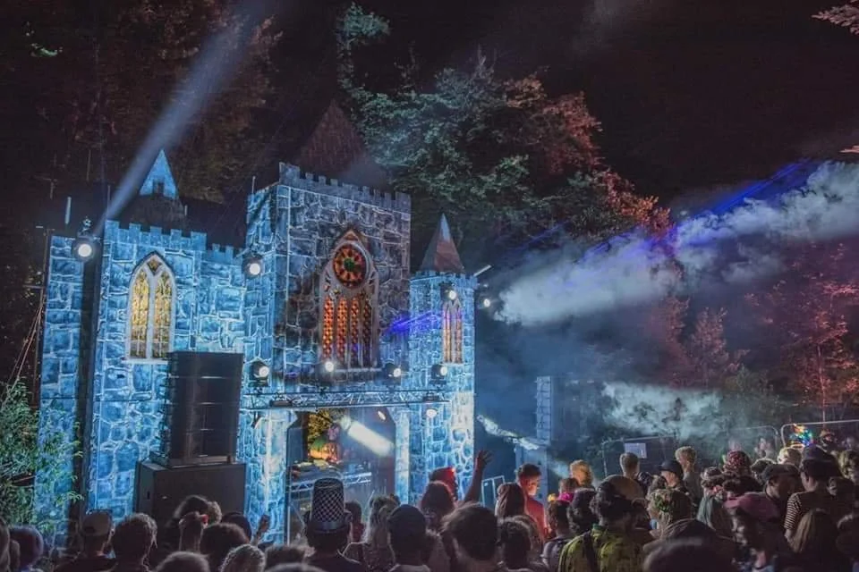 Crowd gathered in front of a castle-shaped stage at nighttime with colorful lighting and smoke effects during a festival or concert.