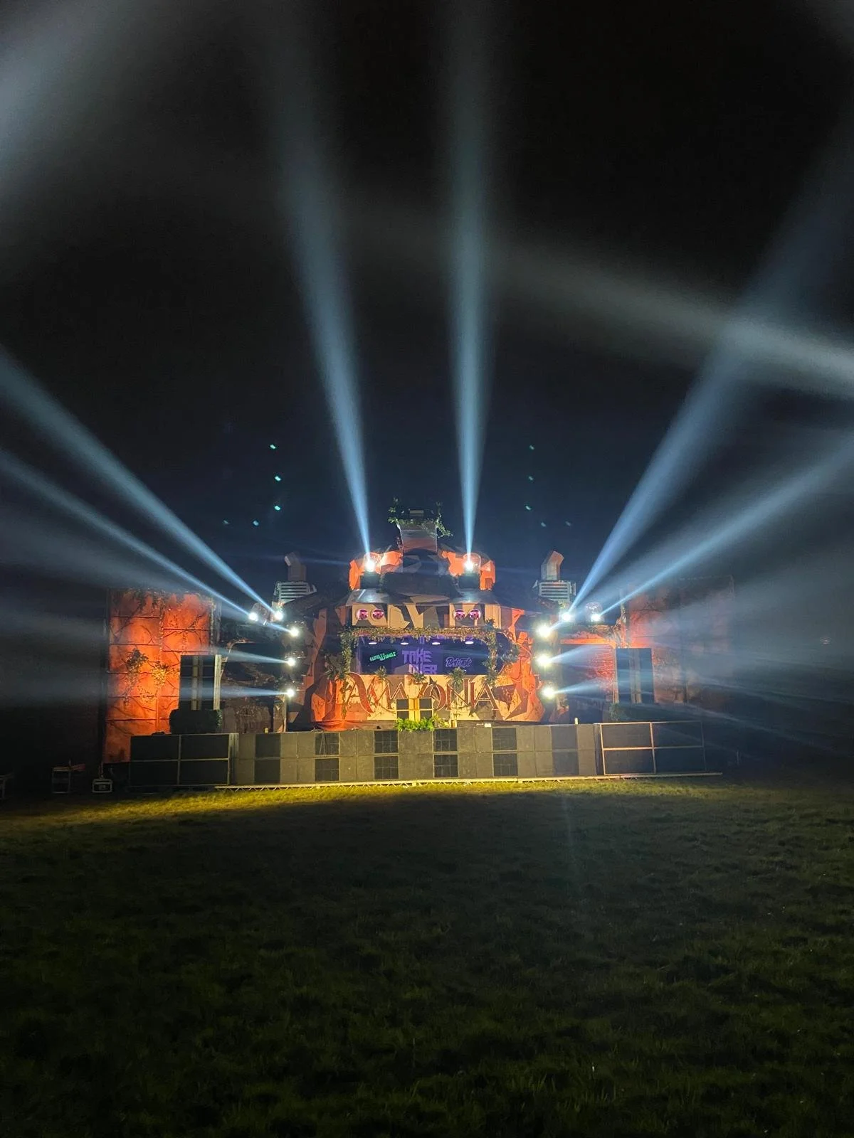 Nighttime outdoor scene with DJ booth or stage, illuminated by bright spotlights and colorful lights, with electronic screens displaying graphics or text, and a grassy area in the foreground.
