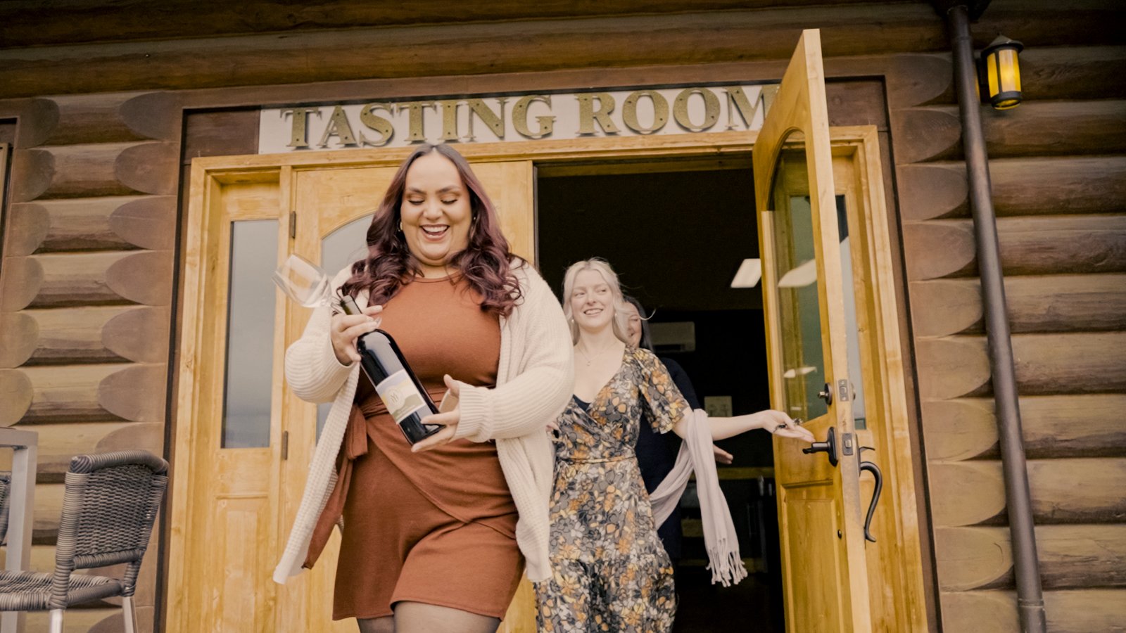 Two women exiting a tasting room, one holding a wine bottle and glass.