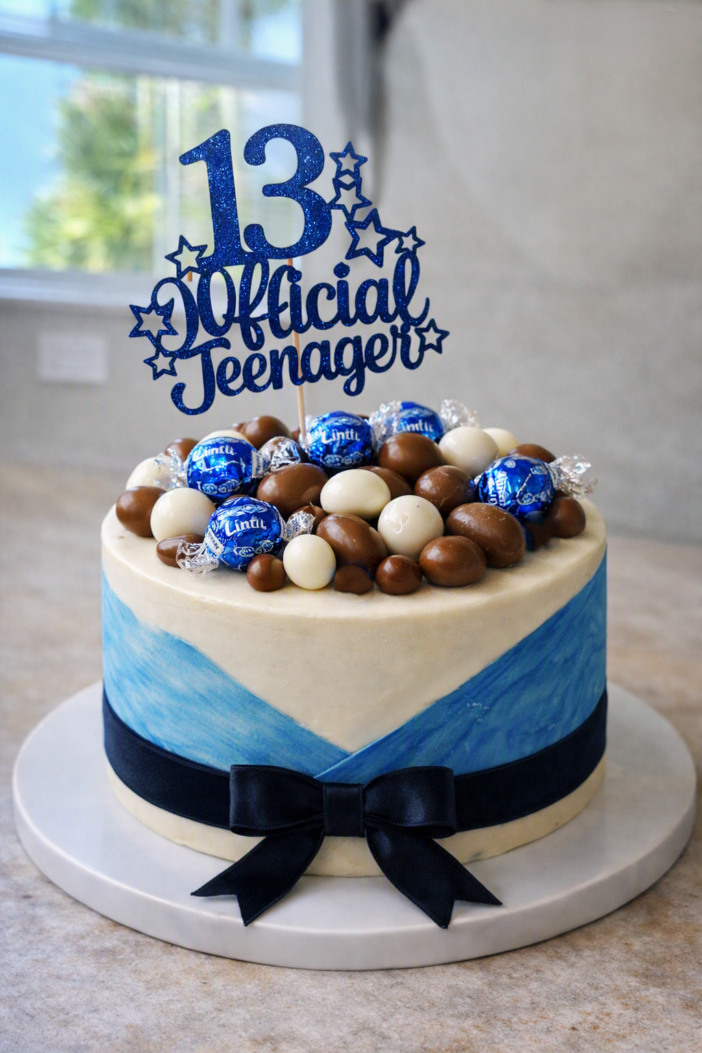 Signature vanilla sponge, filled with Nutella, covered in rich chocolate ganache, and wrapped in smooth fondant with a deep blue velvet ribbon. The top is decorated with blue fondant accents, chocolate‑covered almonds, and blue Lindt chocolates.