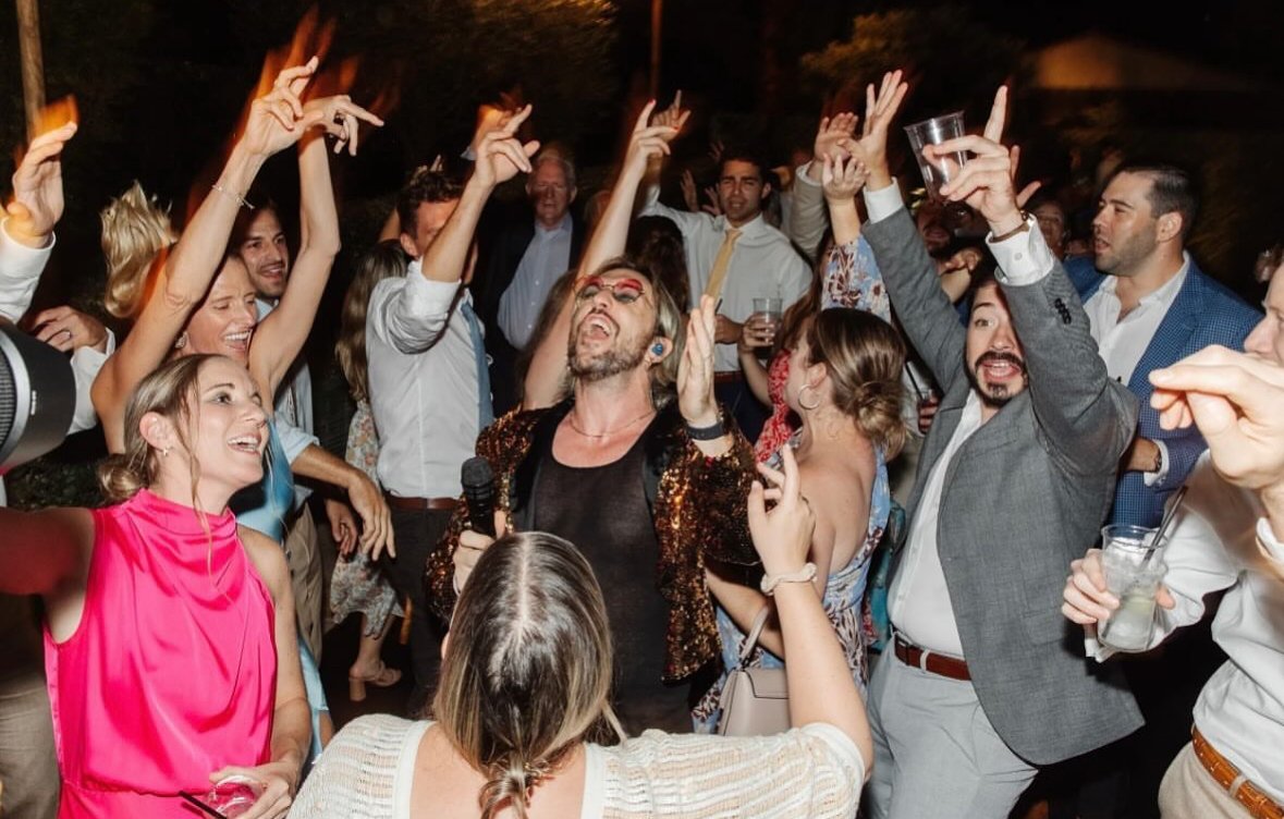 A lively party scene with people dancing and singing, featuring a man holding a microphone. The crowd is happy, with arms raised and drinks in hand, indicating a joyful celebration.
