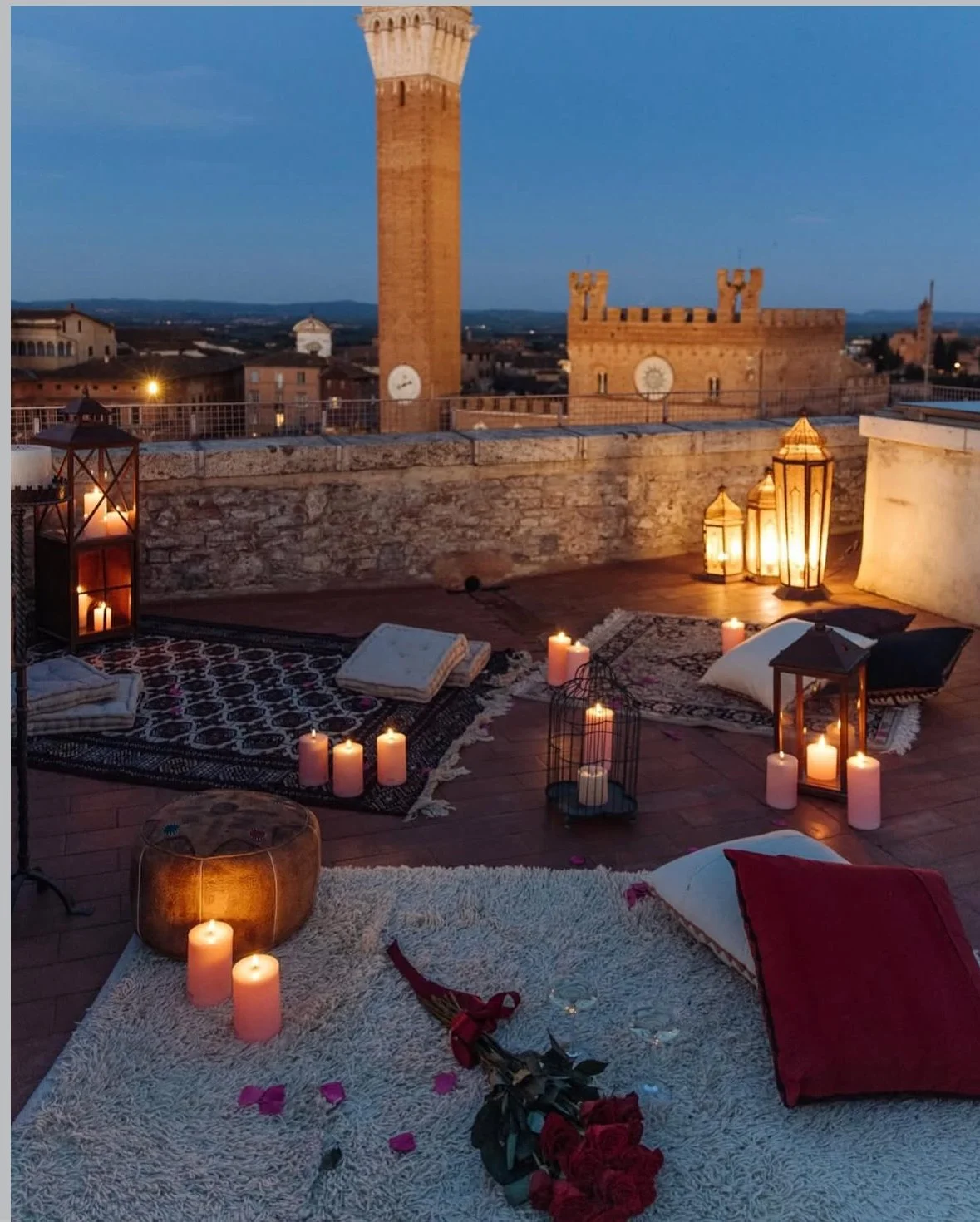 Rooftop evening setting with candles, lanterns, blankets, and pillows, overlooking historical towers and a cityscape.