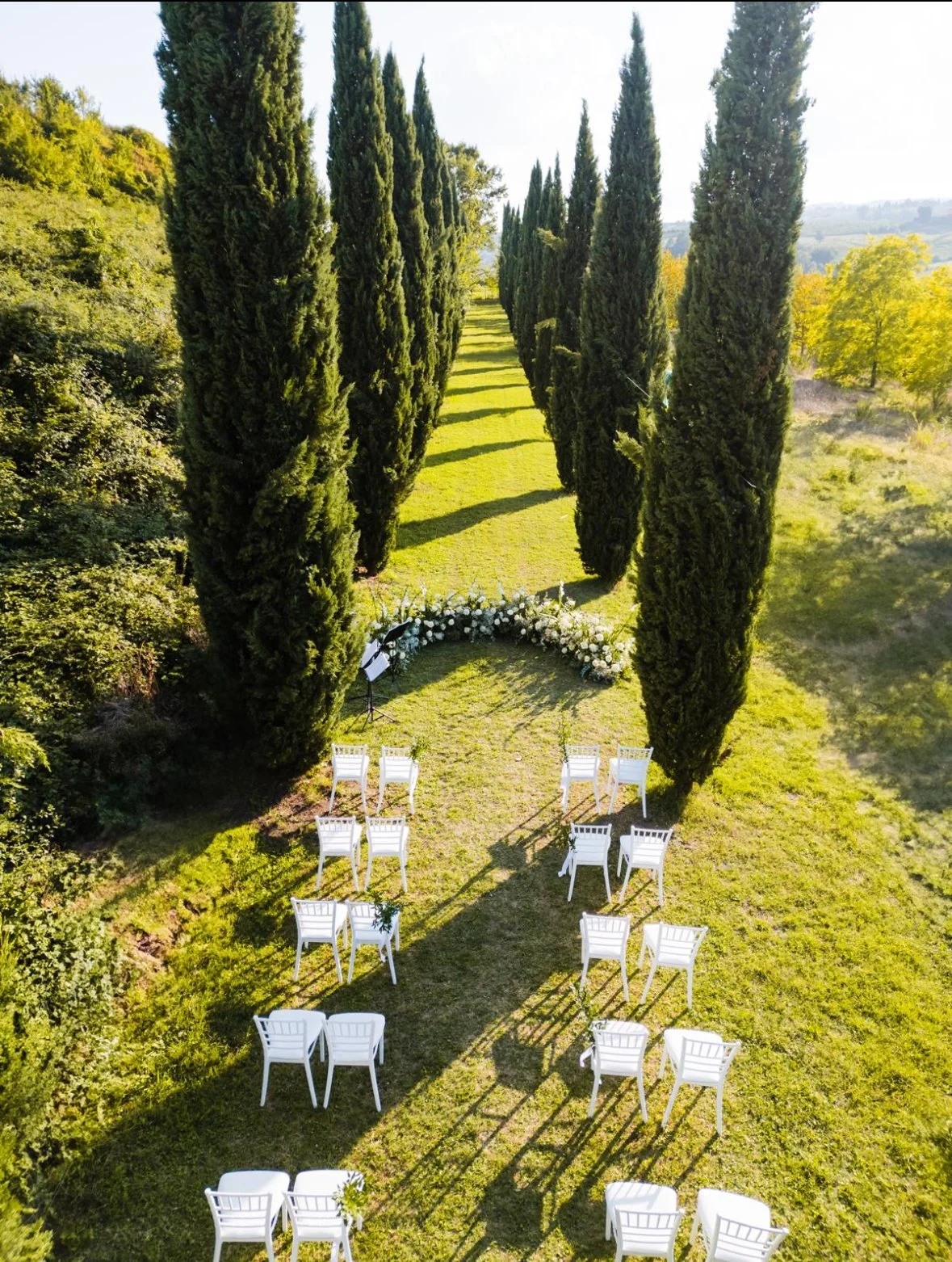 Outdoor wedding setup with white chairs arranged in rows on a grassy aisle lined with tall evergreen trees. Floral arrangements and music stand at the back.